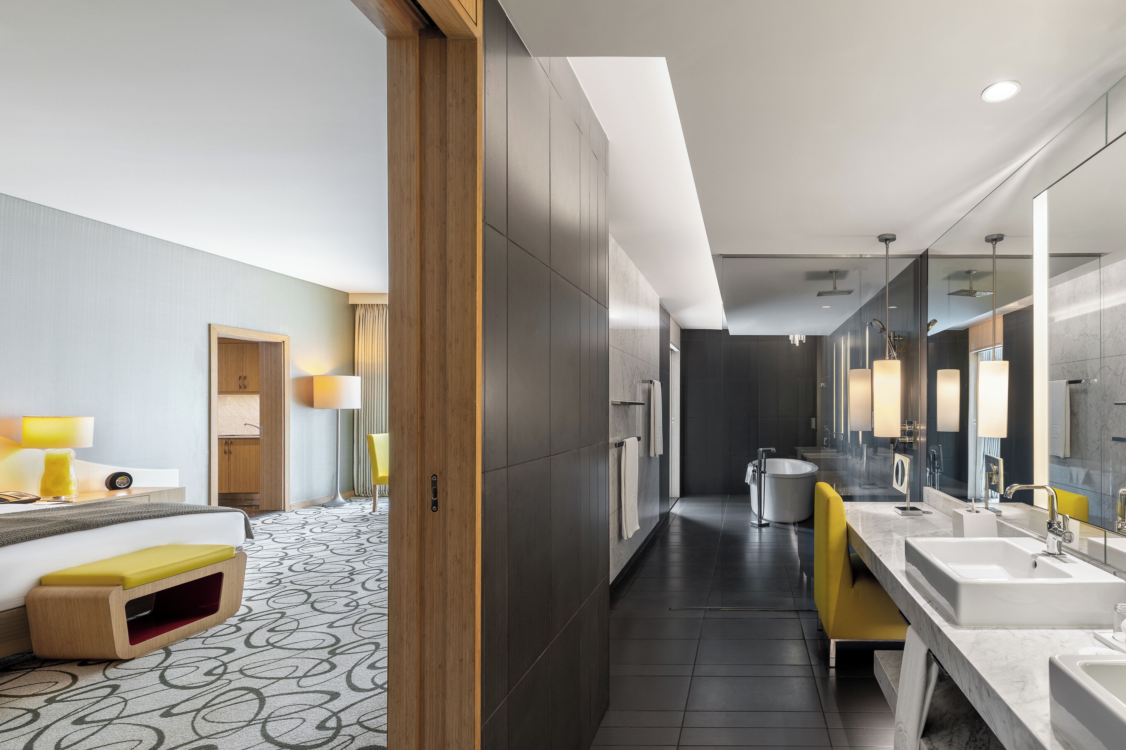 A modern hotel bathroom with a large mirror, dual sinks, a bathtub, and a yellow chair.