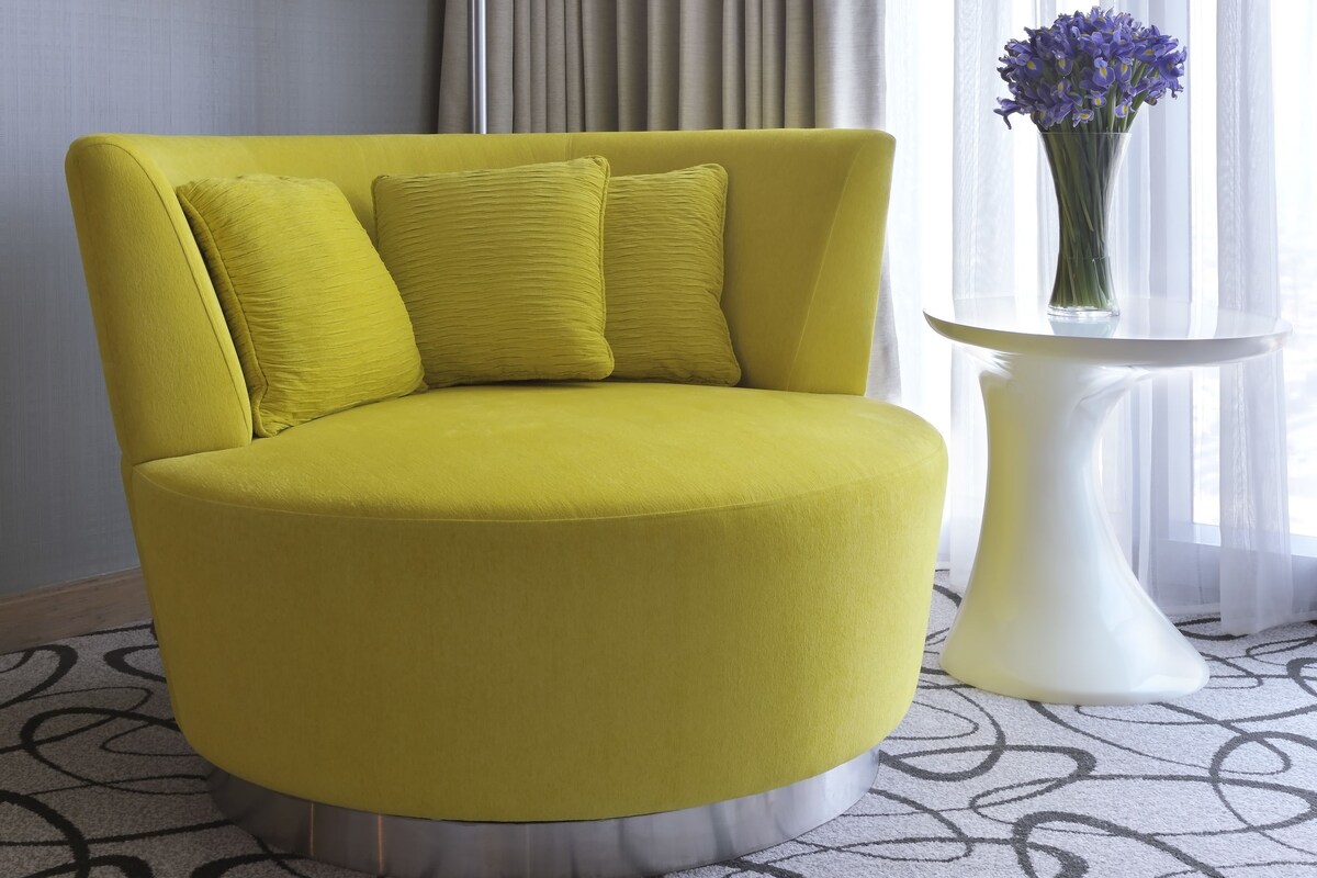 A yellow armchair with two cushions, a white floor lamp, a small white table with a vase of flowers, and a patterned carpet.