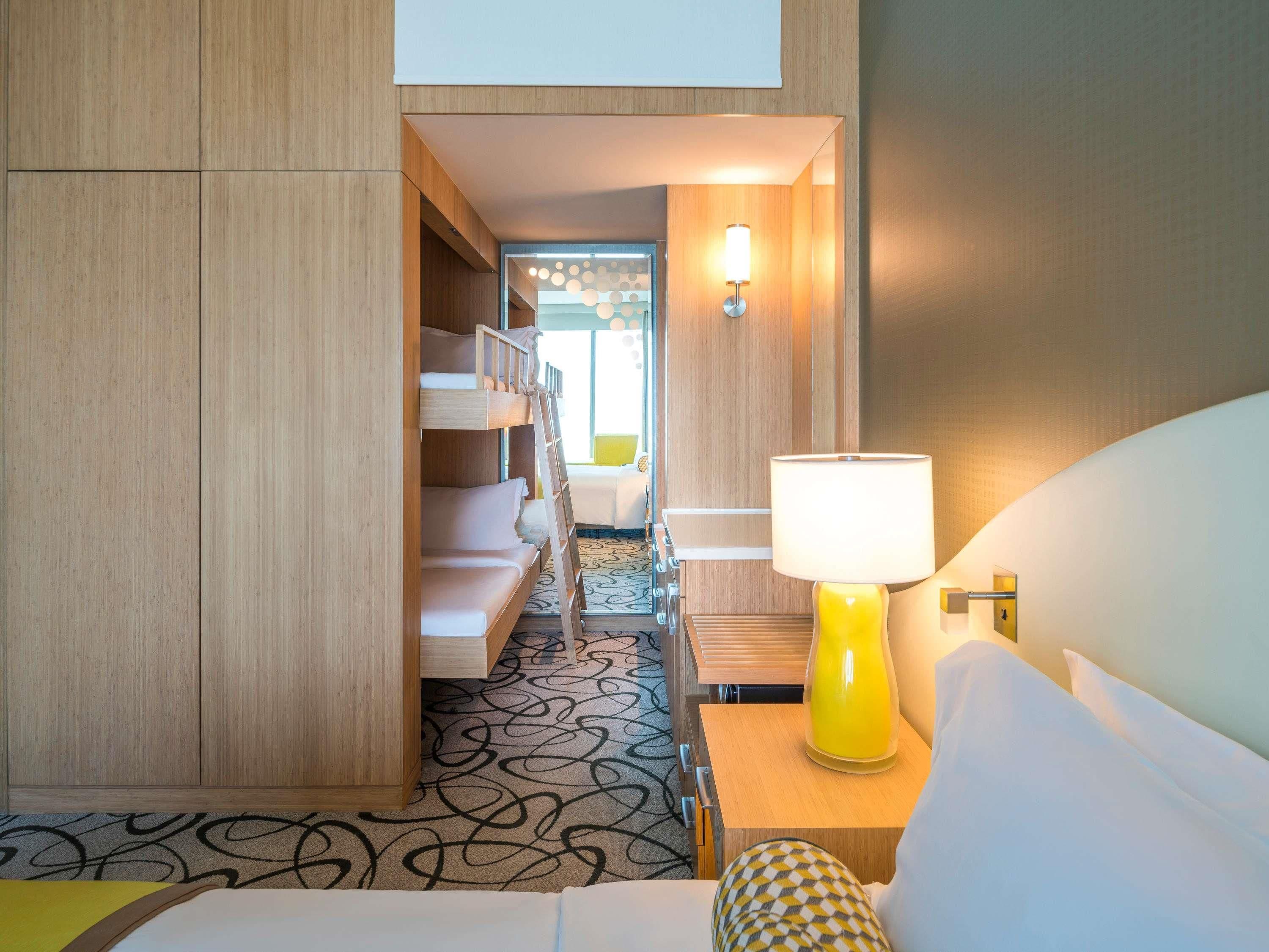 Quarto de hotel com beliches, luminária de cabeceira e carpete estampado.