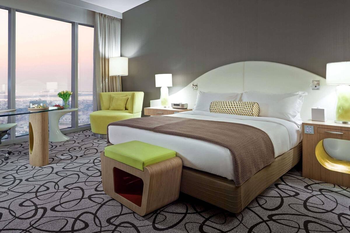 A hotel room with a large bed, a chair, a desk, and a view of the city.