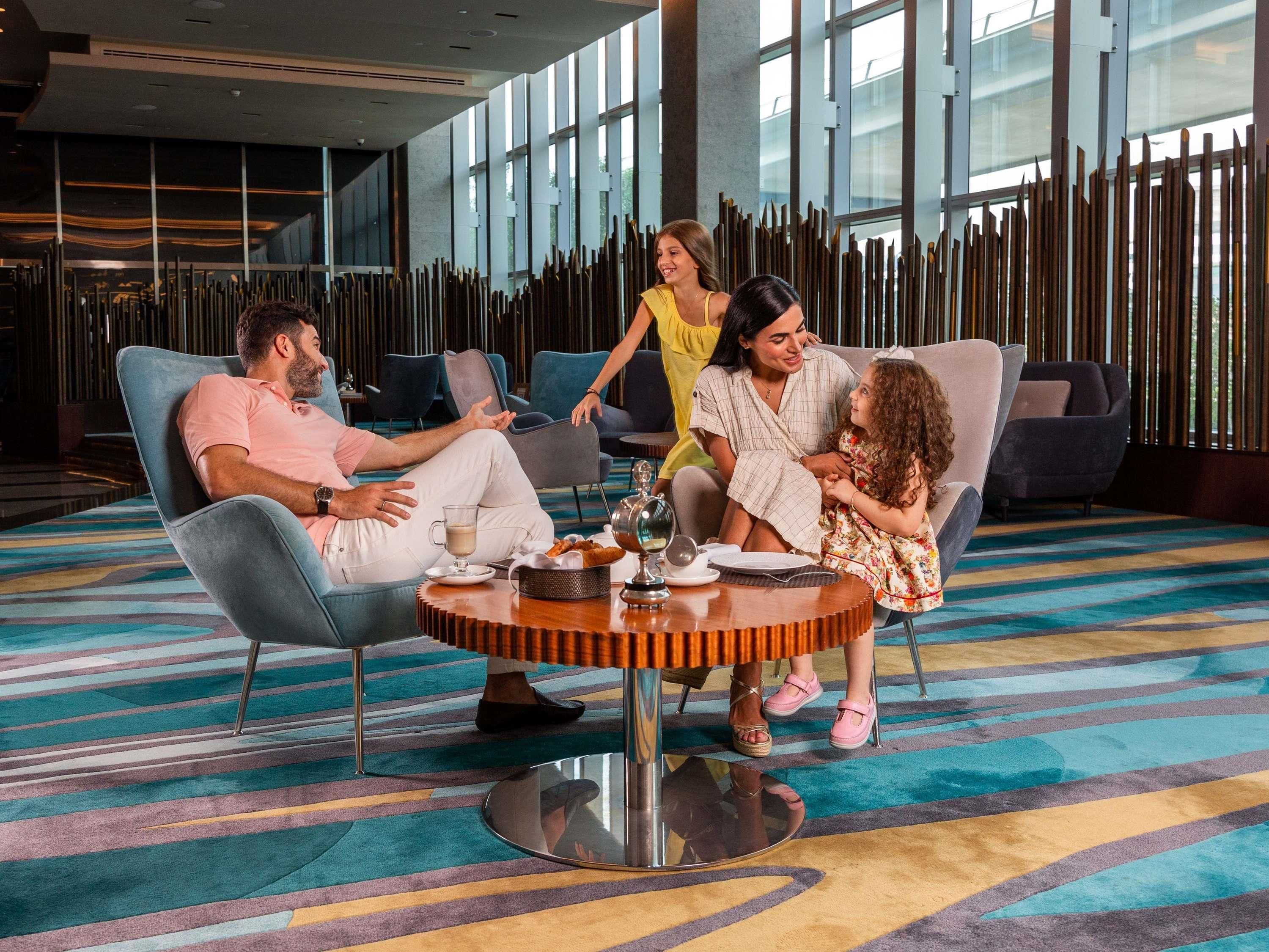 Uma família de quatro pessoas sentada em volta de uma mesa redonda em um lobby moderno de hotel, com carpete estampado e ripas de madeira verticais ao fundo.