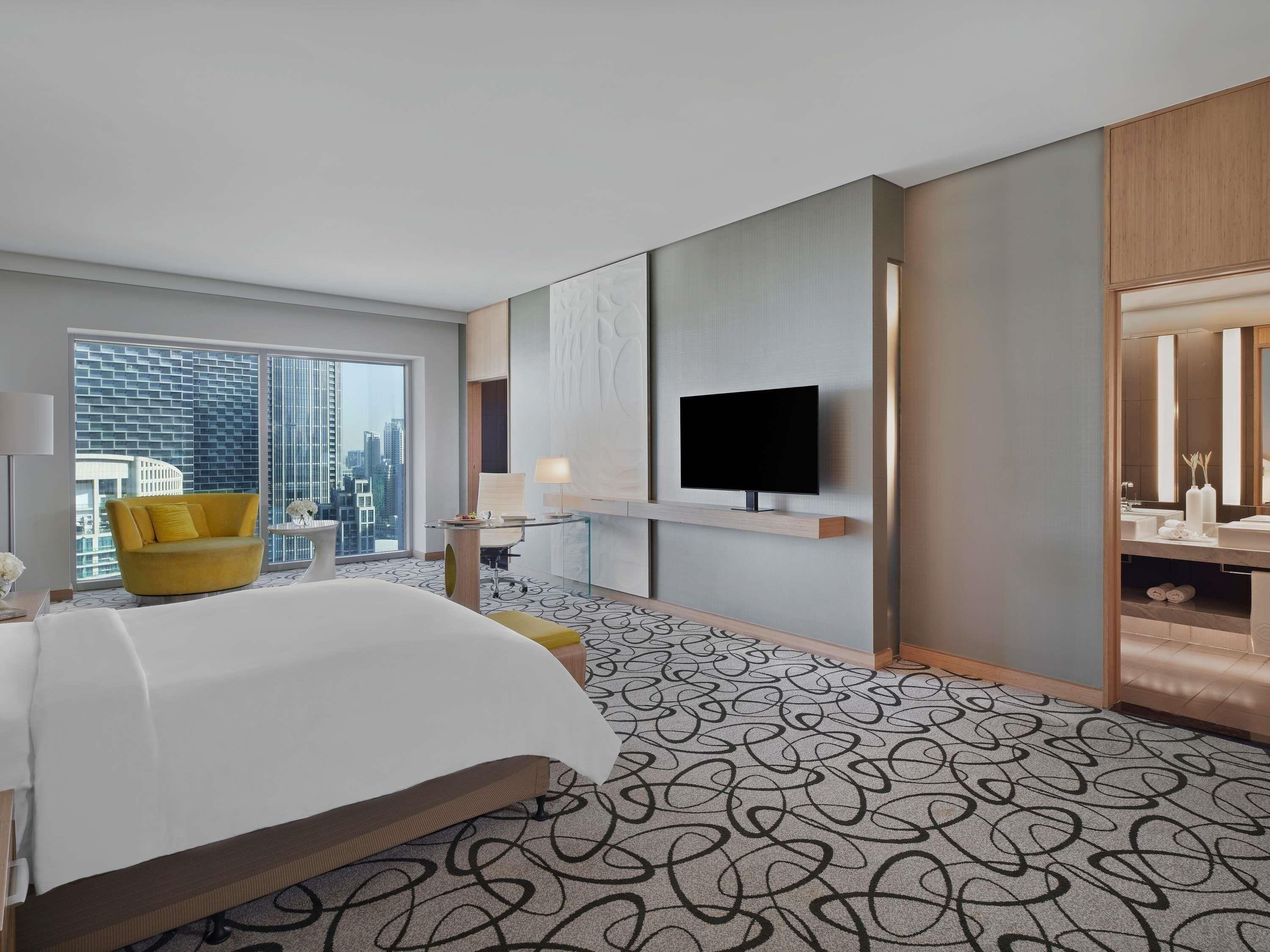 A hotel room with a bed, a yellow armchair, a TV, a large window, and a city view.