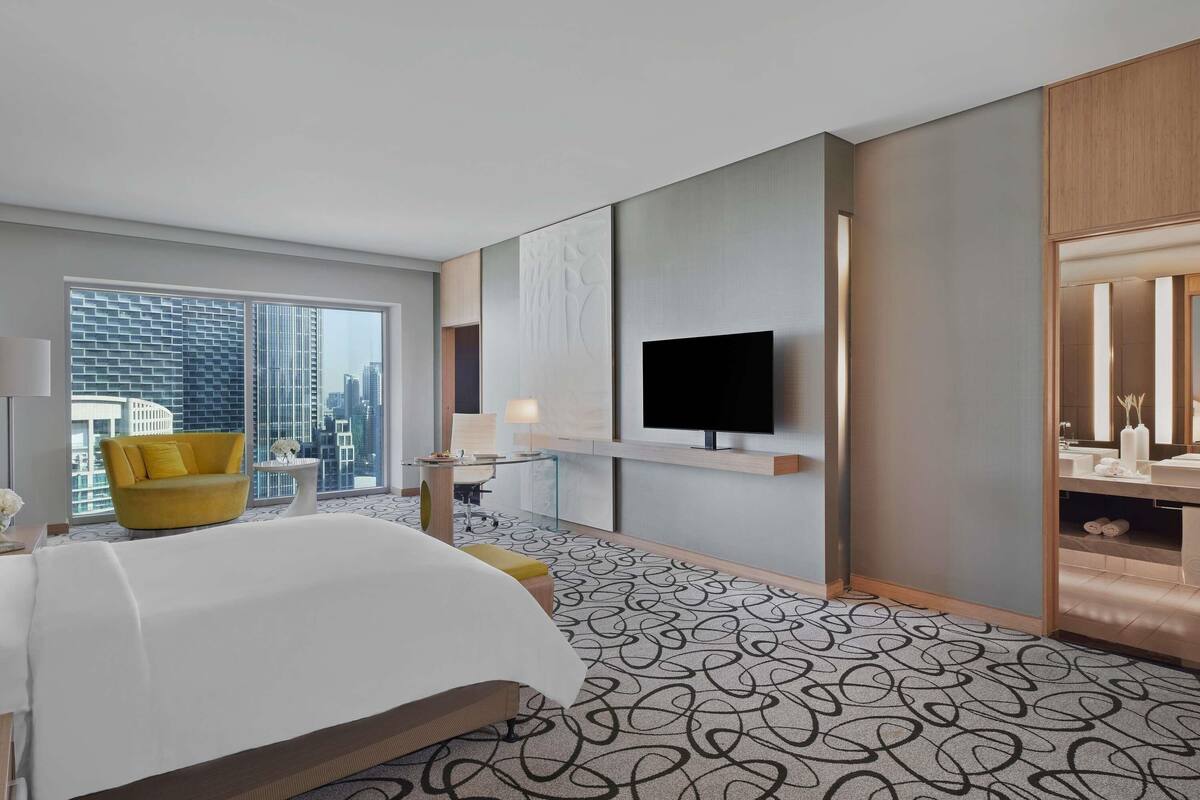 A hotel room with a bed, a yellow armchair, a TV, a large window, and a city view.