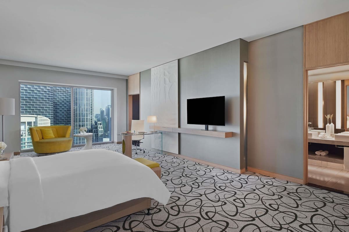 A hotel room with a bed, a yellow armchair, a TV, a large window, and a city view.