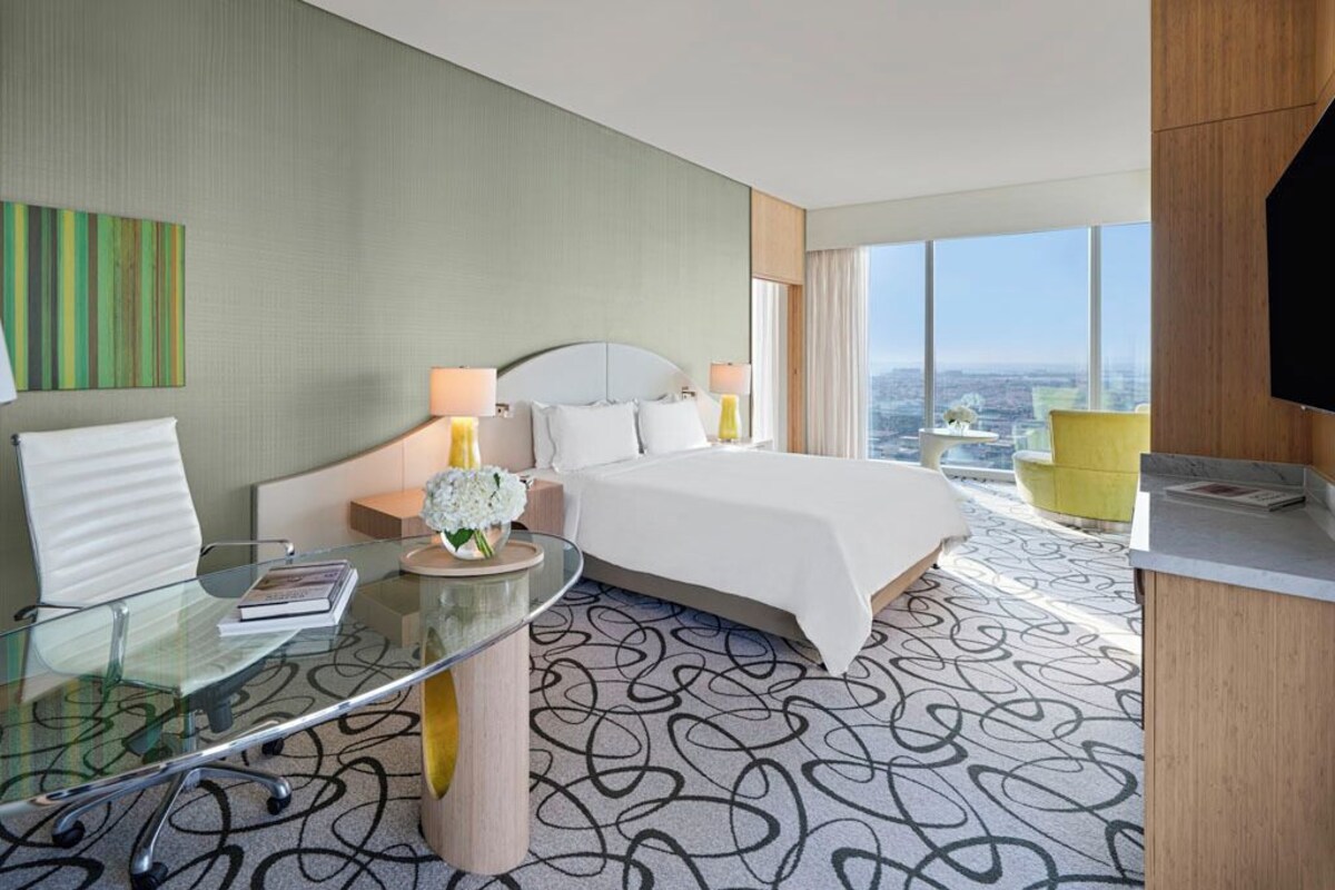 A modern hotel room with a large bed, a glass desk, a view of the city, and a television.
