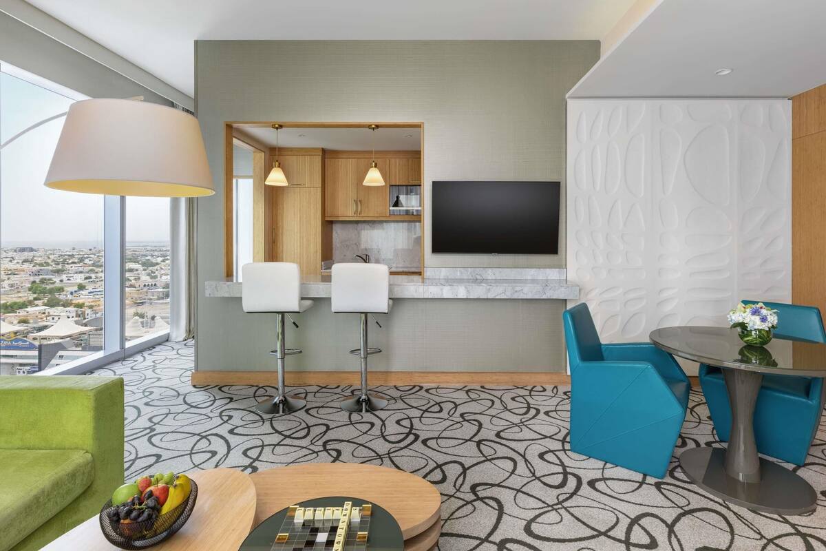 A modern hotel room with a large window, a flat-screen TV, a dining area with blue chairs, and a green sofa.
