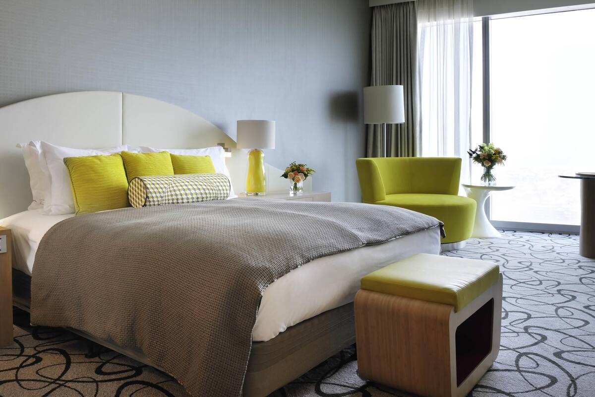 A hotel room with a large bed, a bright yellow armchair, a small table with a vase, and a view of the outdoors.