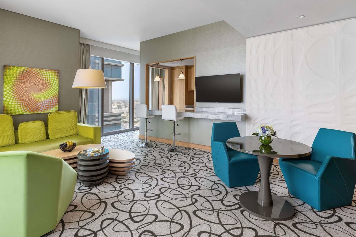 A hotel room with a sofa, chairs, a table, a TV, and a large window with a city view.