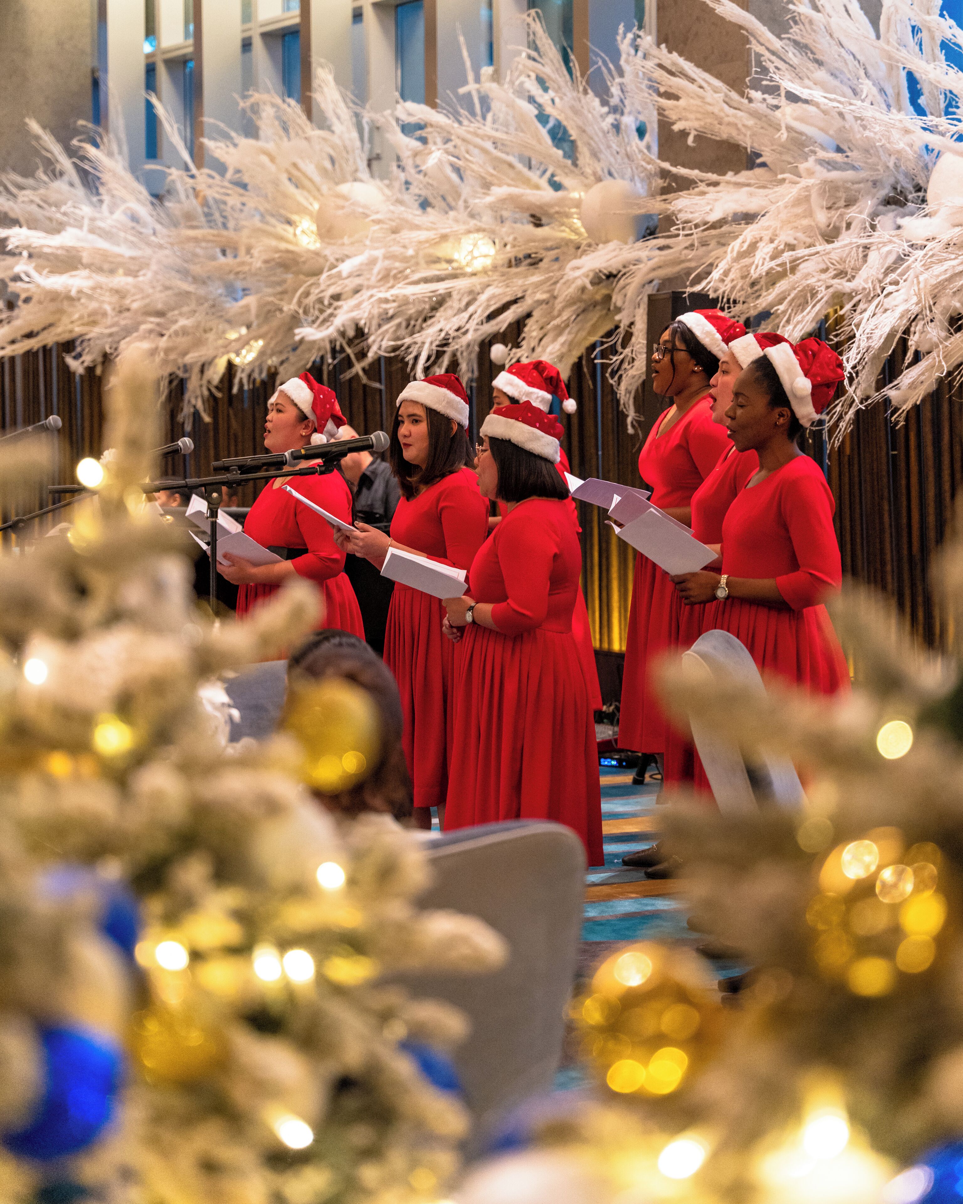 A group of people in red dresses and Santa hats singing in a decorated indoor space.