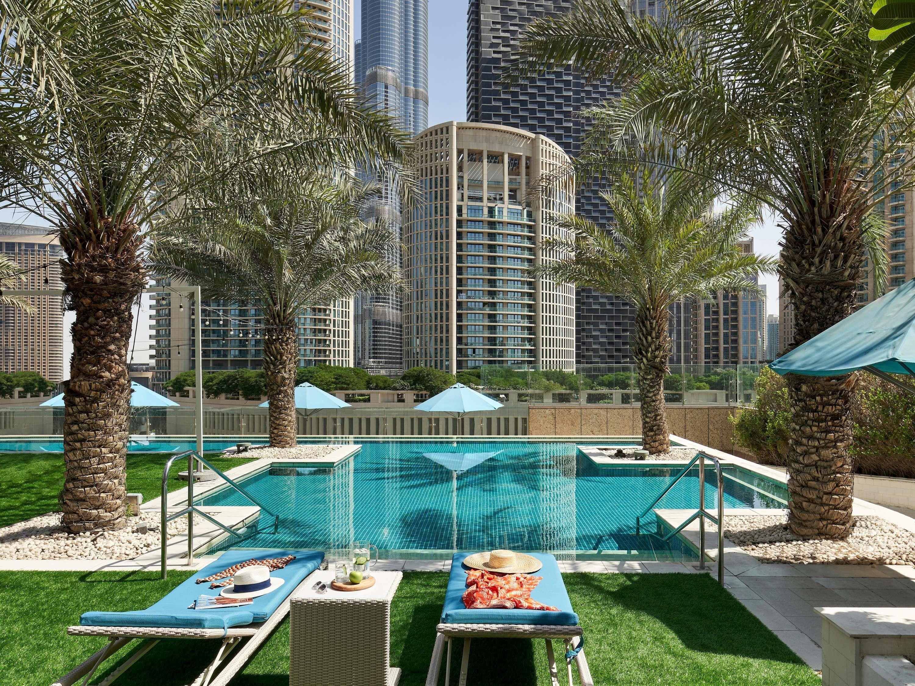 A pool area with lounge chairs and umbrellas, surrounded by palm trees and modern high-rise buildings in the background.