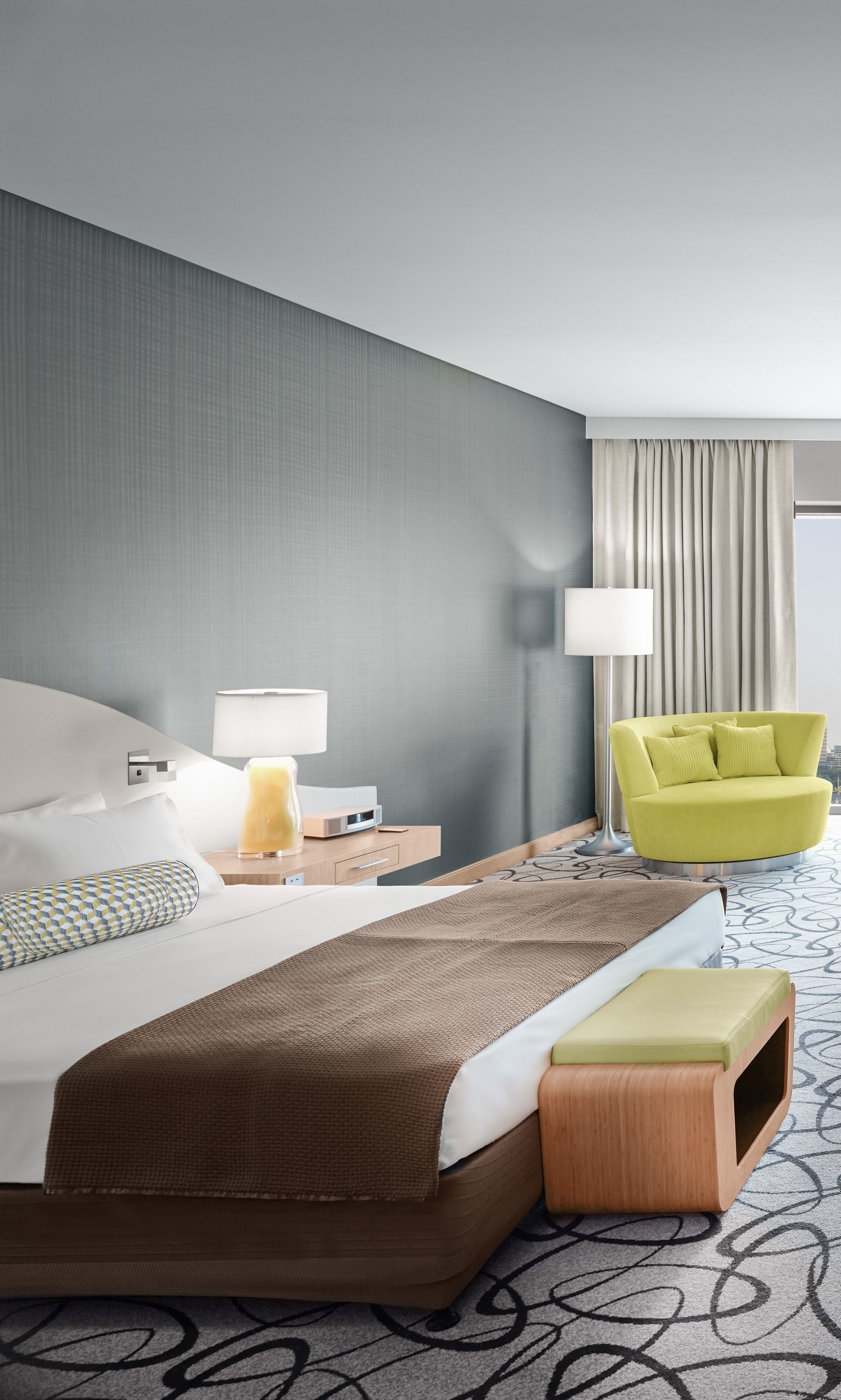 Ein Hotelzimmer mit Bett, Nachttischen, einer Lampe, einem Sessel und Blick auf die Stadt.