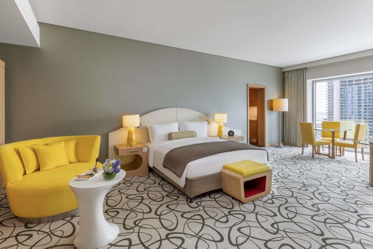 A hotel room with a bed, a yellow armchair, a small table with a lamp, a red storage box, and a view of a cityscape through a large window.