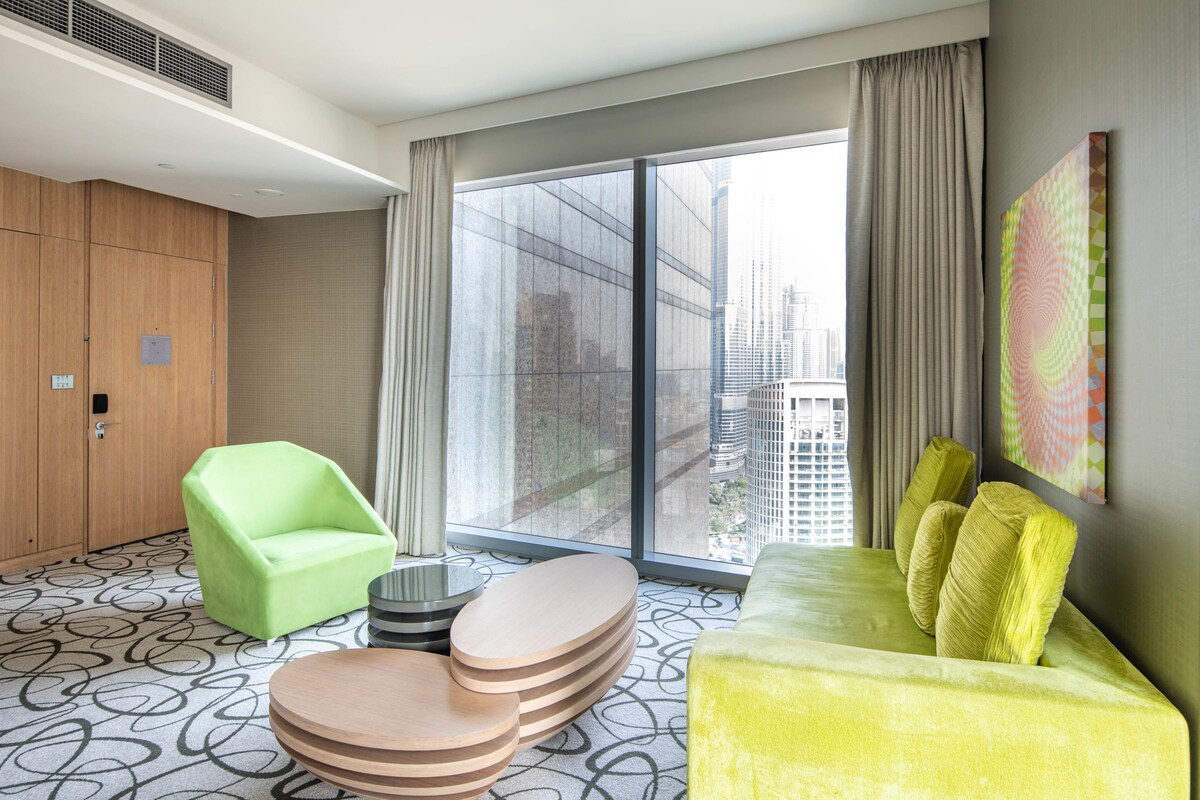 A modern hotel room with a large window, a green chair, a yellow sofa, and a unique patterned floor.
