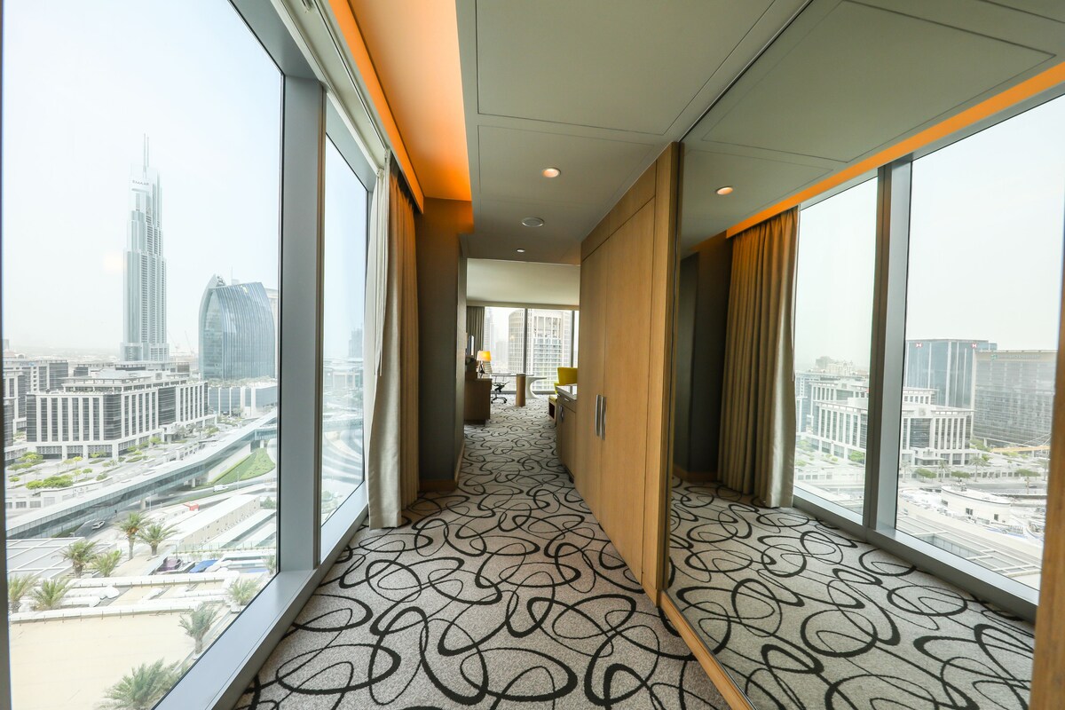 A hotel corridor with a patterned carpet, large windows offering city views, and a wooden door.