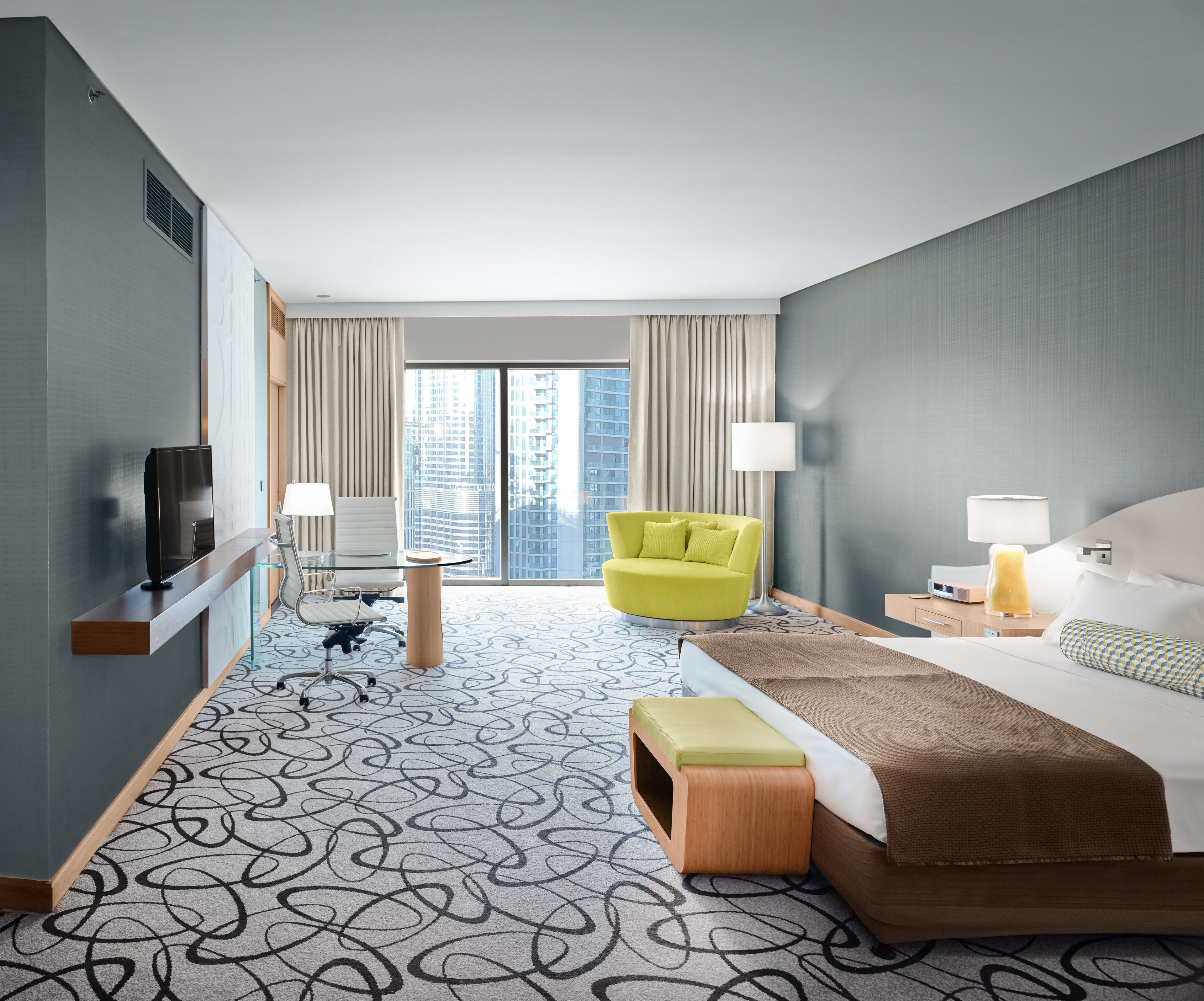 Habitación de hotel moderna con una cama grande, un sillón amarillo brillante, una mesa de vidrio con una silla, un televisor en un estante de madera y vistas de edificios altos por la ventana.