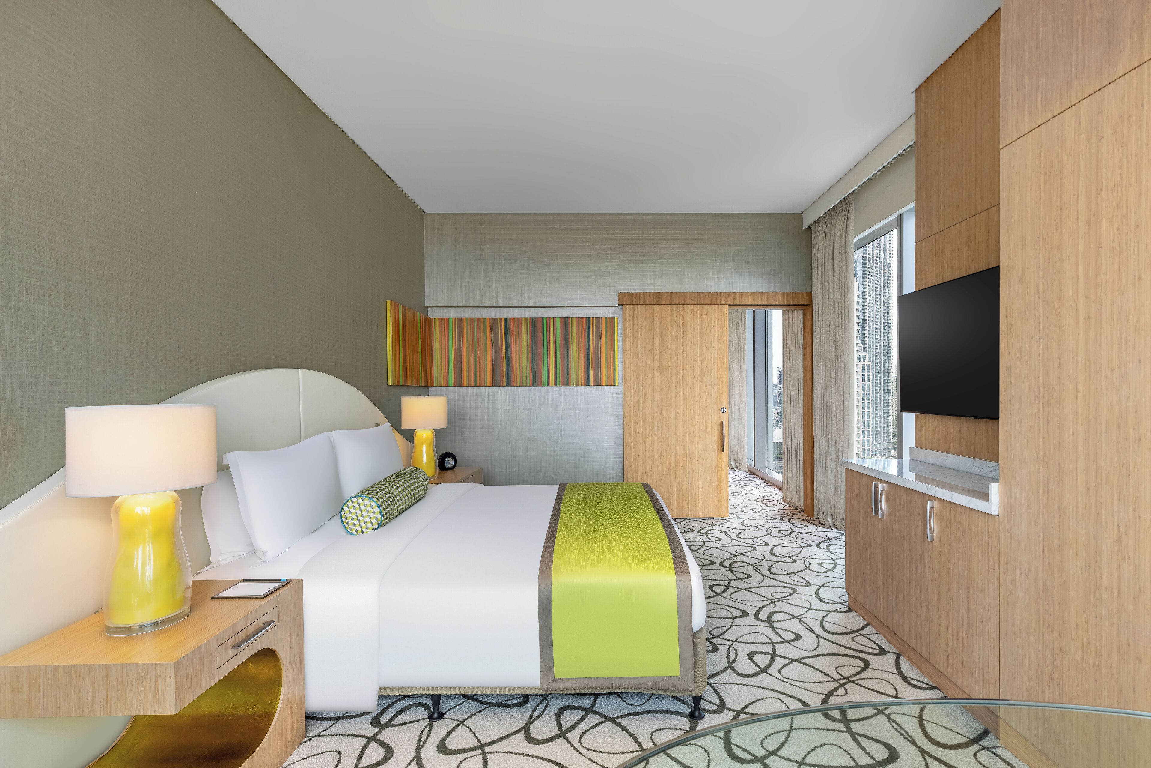 Habitación de hotel con cama, lámparas de noche, televisor, ventana y alfombra estampada.