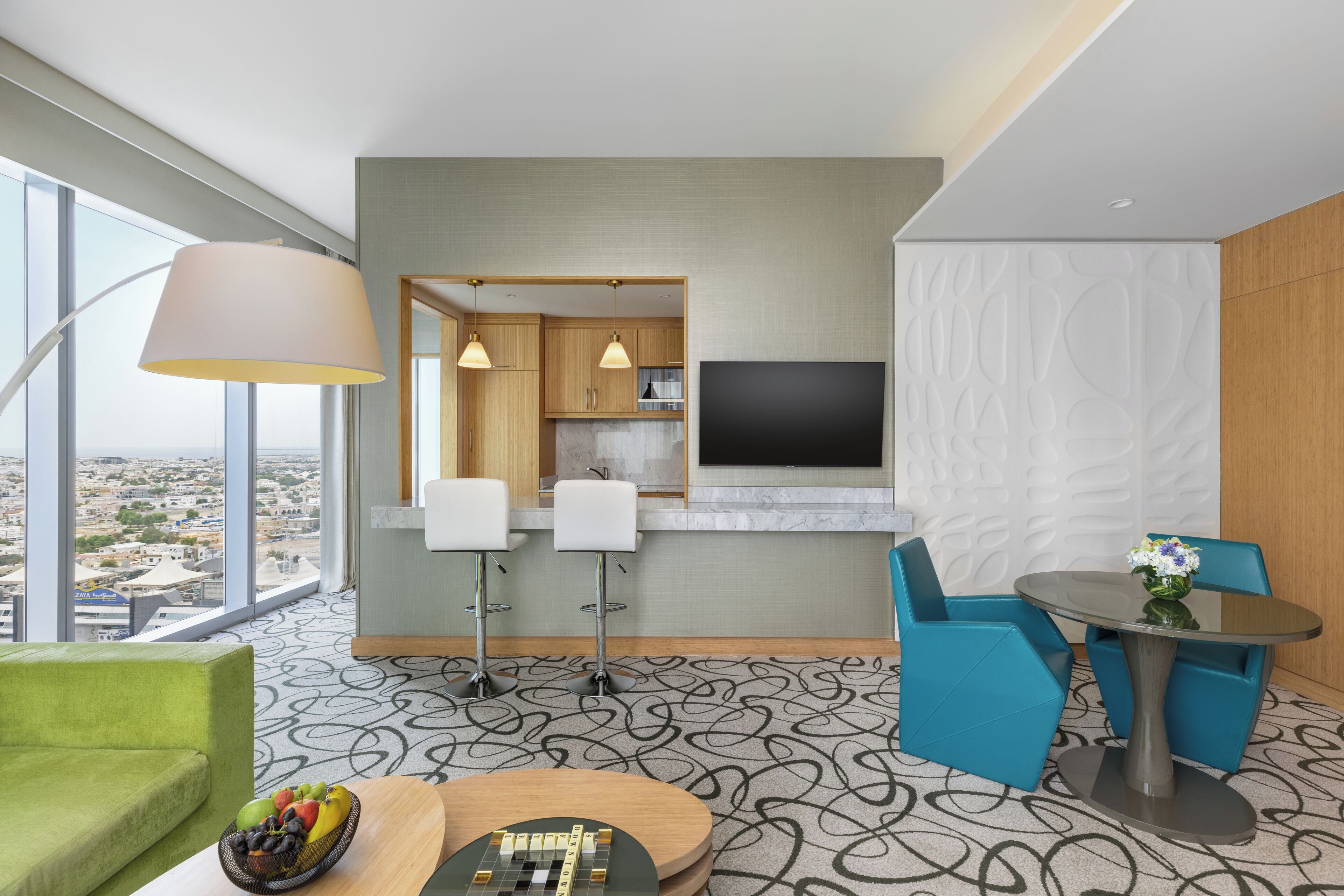 Une chambre d’hôtel moderne dotée d’une grande fenêtre, d’un coin repas avec des chaises bleues, d’un coin cuisine et d’un téléviseur à écran plat fixé au mur.