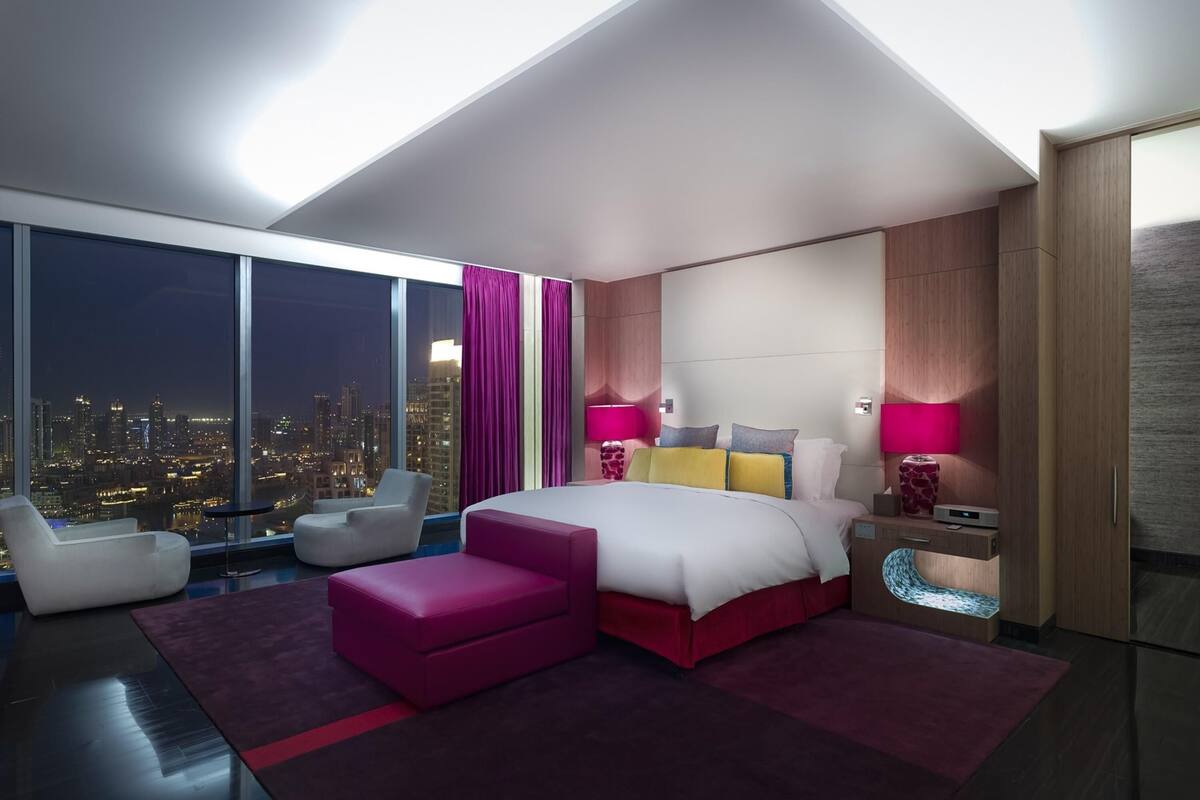 A modern hotel room with a large bed, a city view, a desk with a lamp, and a seating area.