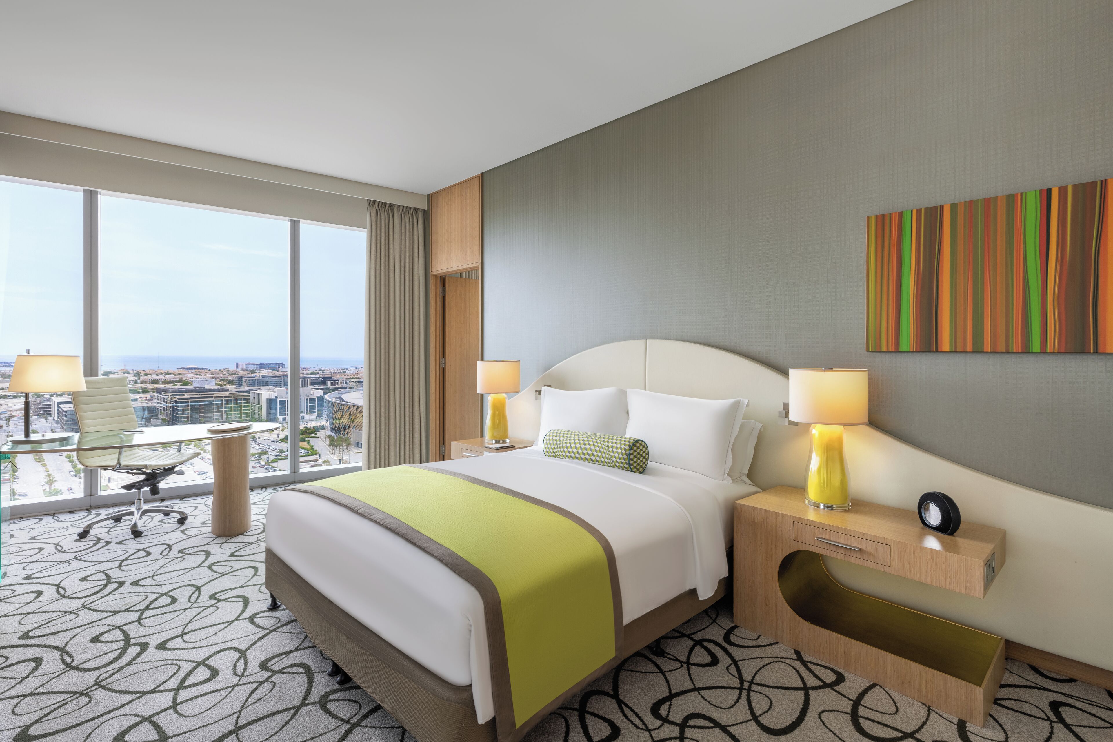 Quarto de hotel com uma cama grande, duas mesas de cabeceira, uma escrivaninha e uma cadeira, e vista para a cidade.