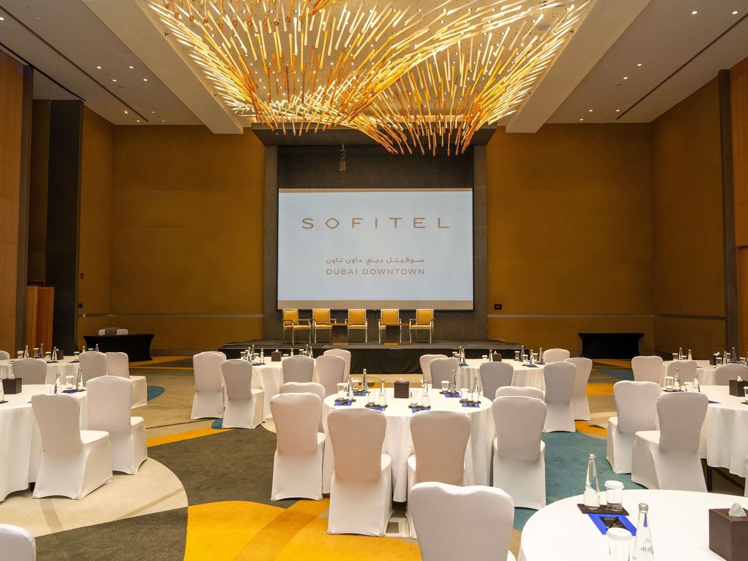 Un salón de conferencias con sillas y mesas blancas, una pantalla grande que muestra 'SOFITEL DUBAI DOWNTOWN' y una lámpara de techo de diseño único.