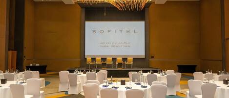 Un salón de conferencias con sillas y mesas blancas, una pantalla grande que muestra 'SOFITEL DUBAI DOWNTOWN' y una lámpara de techo de diseño único.