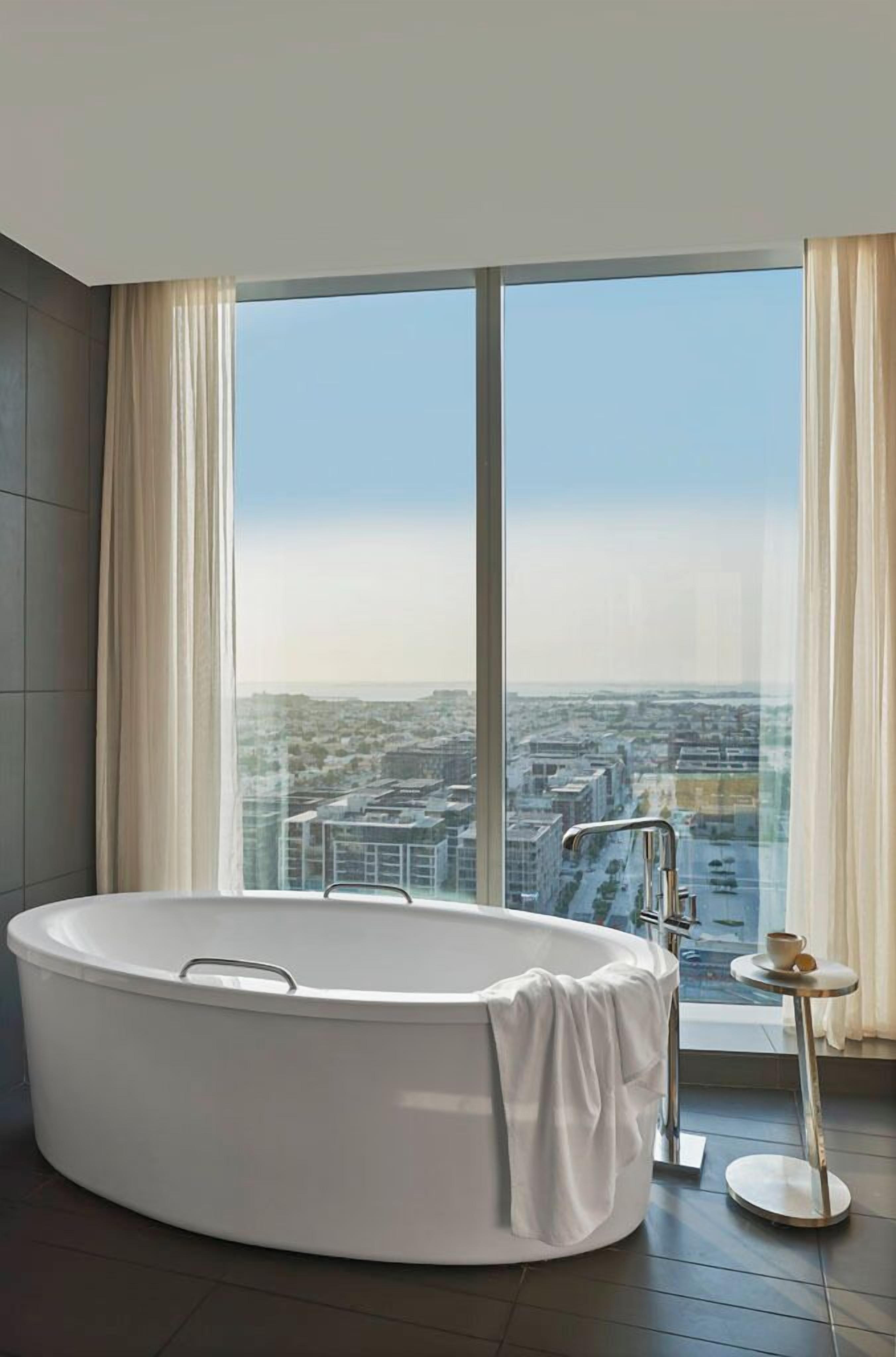 A modern bathroom with a large bathtub, a view of a cityscape through floor-to-ceiling windows, and a small round table with a cup.