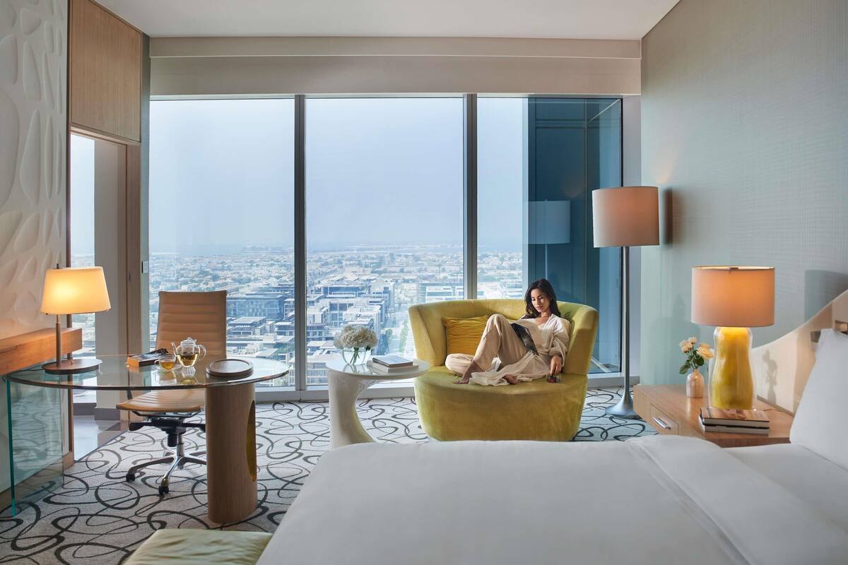 A hotel room with a large bed, a yellow armchair, a glass table with a lamp, and a view of a cityscape through large windows.