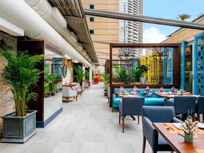 An outdoor dining area with seating and plants, a brick wall, and a tall building in the background.