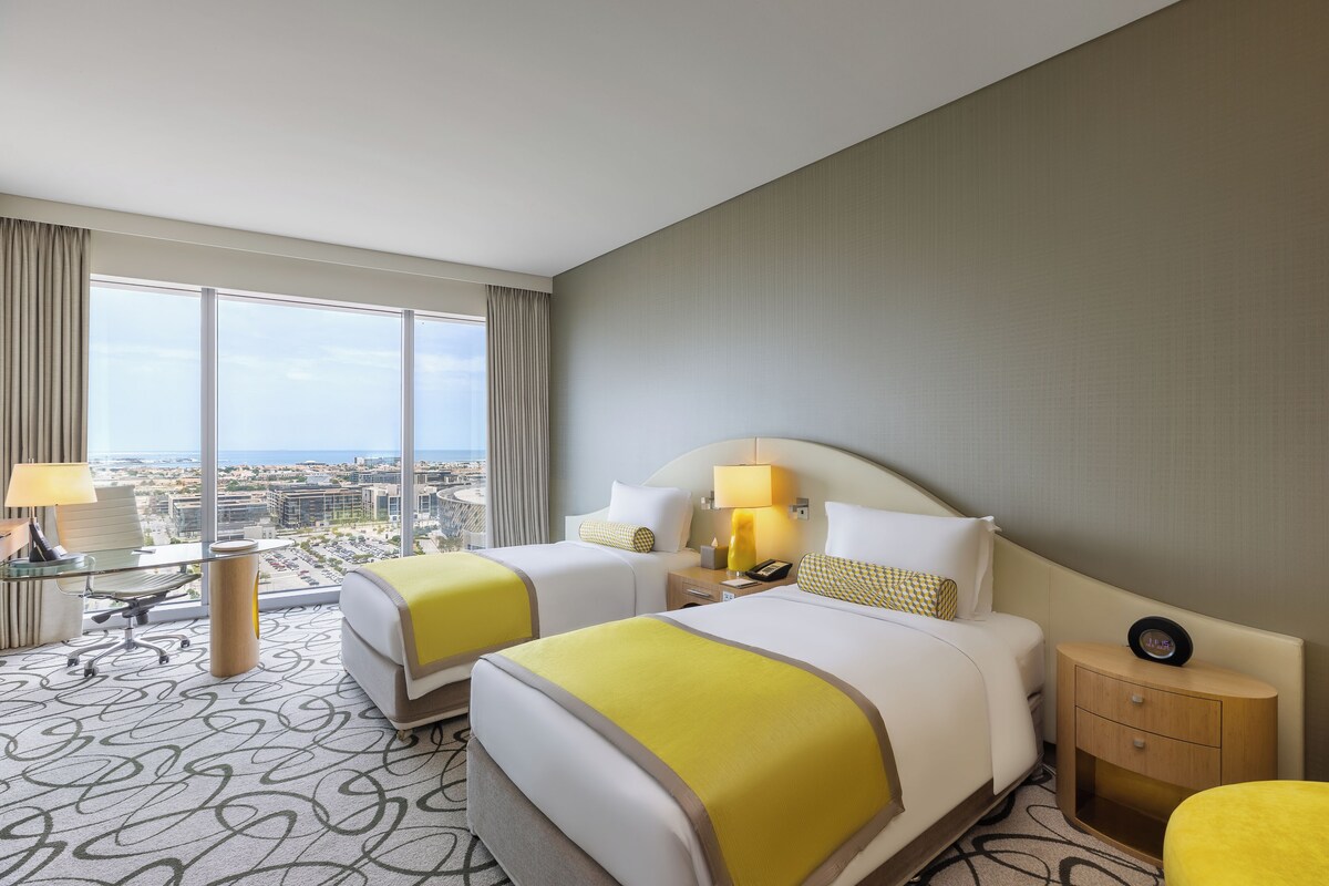 A hotel room with two beds, a desk, and a large window offering a city view.