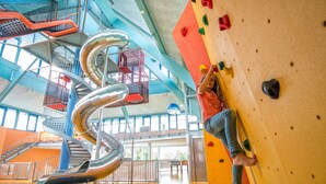A person climbing a rock wall with colorful holds in an indoor facility with a spiral slide and a high ceiling. - JUFA Hotel Jülich im Brückenkopf-Park (Jülich)
