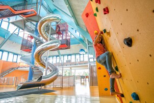 Uma pessoa escalando uma parede de escalada com pontos de apoio coloridos em um espaço interno com um escorregador em espiral e teto alto.