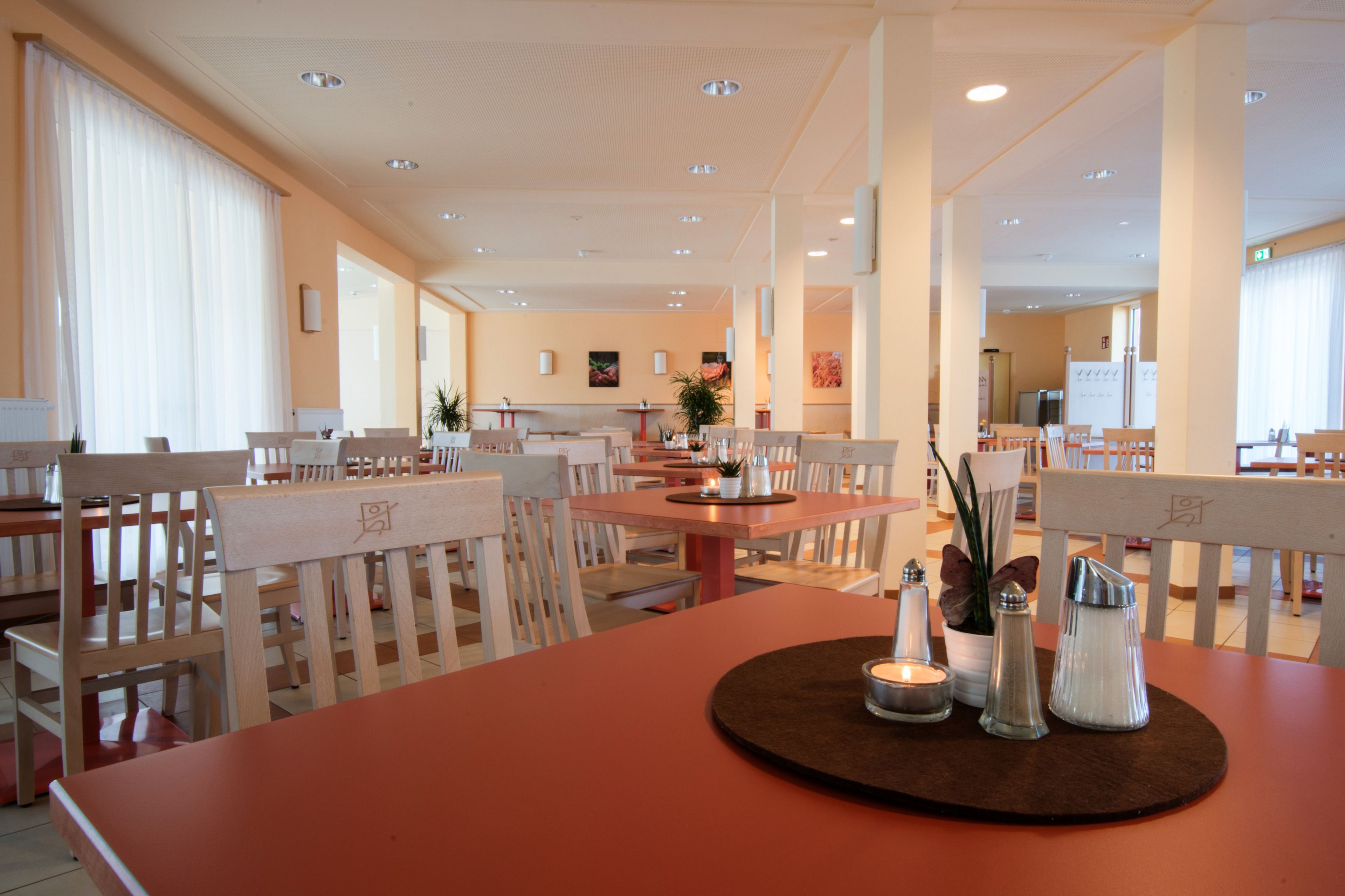 Un comedor amplio con varias mesas y sillas, una mesa central con vela y saleros, y una zona de cocina al fondo.