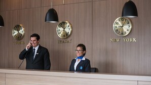 A hotel reception desk with clocks displaying different time zones, a man on the phone, and a woman behind the desk. - Hotel Estação 101 (Itajai)