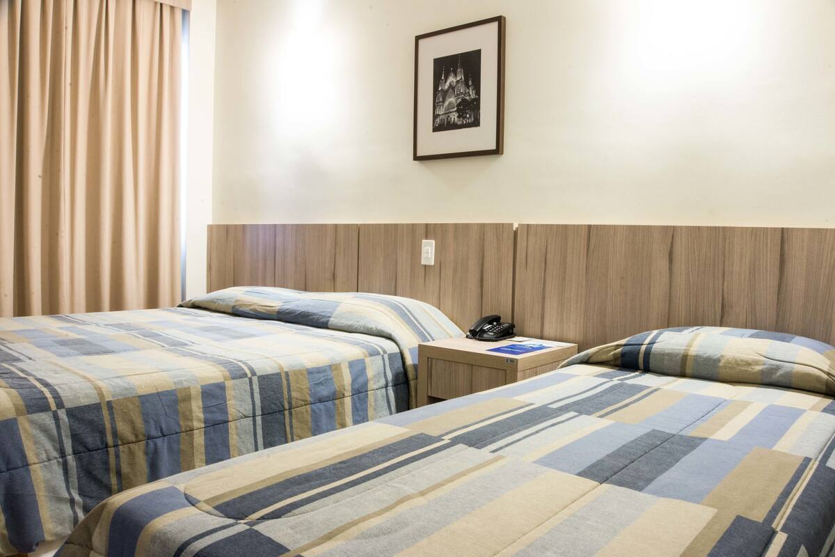 Habitación de hotel con dos camas, una mesita de noche con teléfono, un cuadro en la pared y un cabecero de madera.