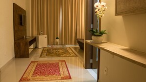 A modern hotel room with a wooden desk, a flat-screen TV, a small refrigerator, a red and beige patterned rug, a potted orchid, and a large window with beige curtains. - Hotel Estação 101 (Itajai)