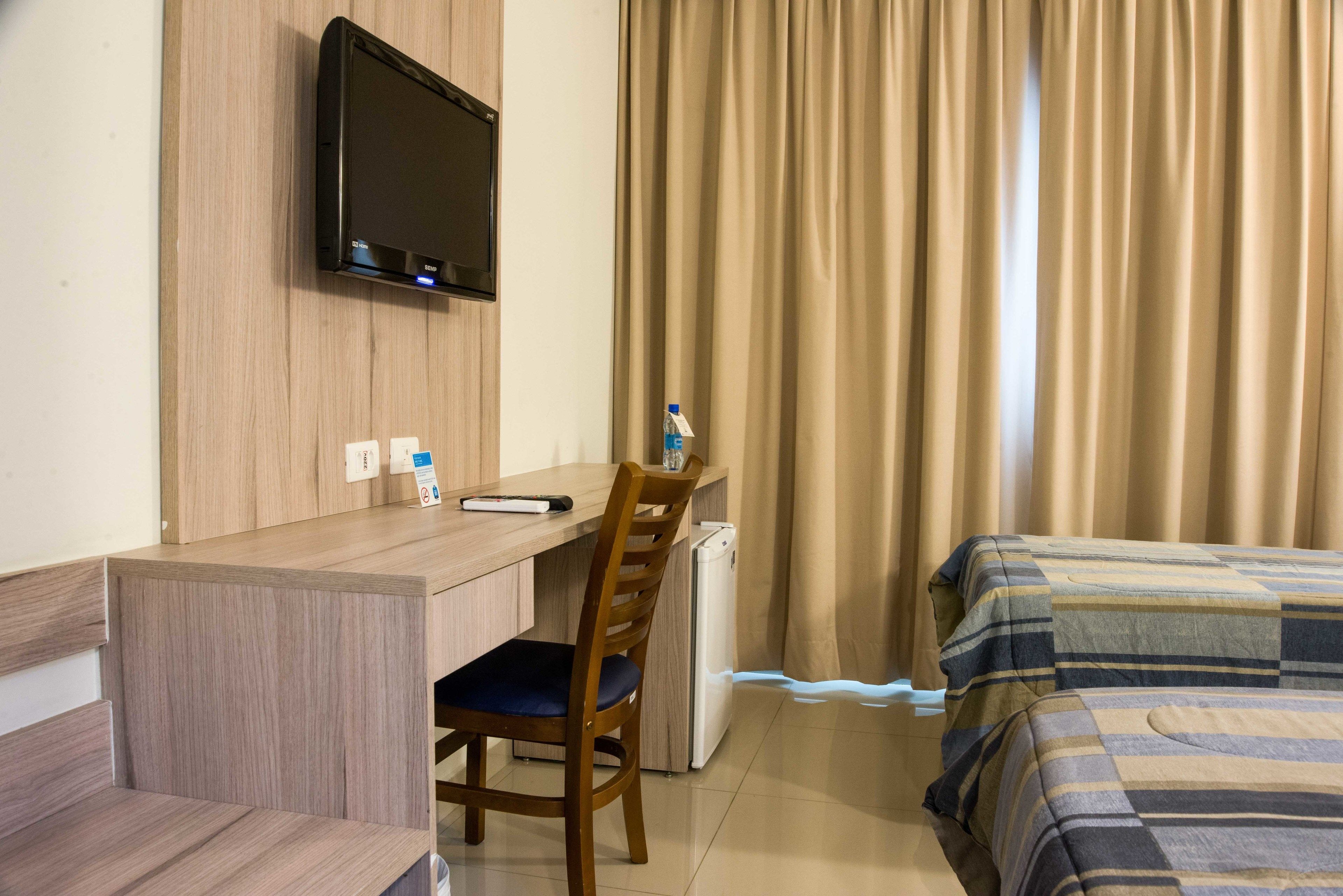 A hotel room with a wooden desk, a chair, a bed with striped bedding, a television mounted on the wall, and a window with beige curtains.