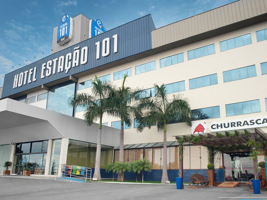A hotel named "Hotel Estação 101" with a modern design, large windows, and a sign for "Churrascaria" indicating a dining option.