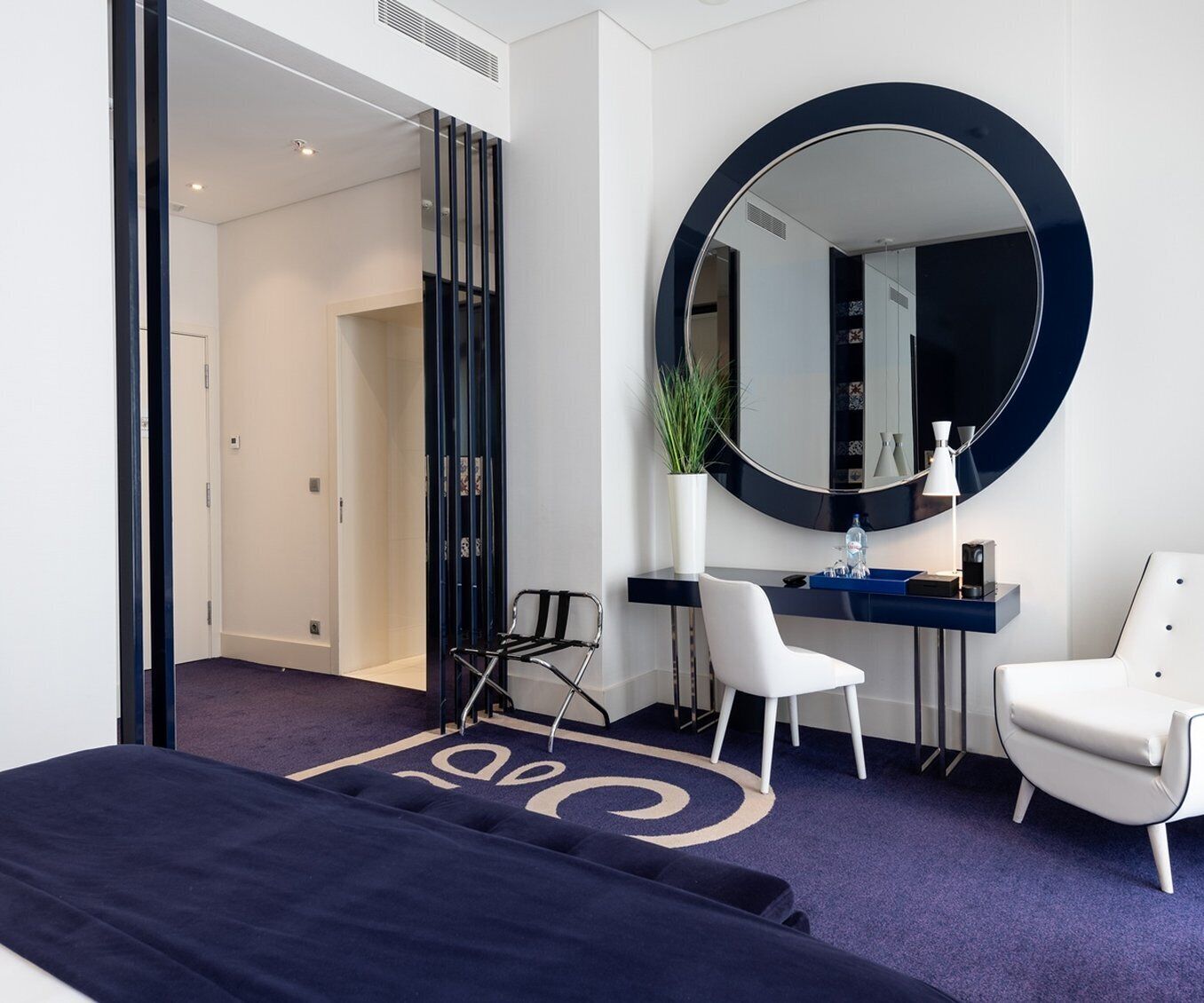 A modern hotel room with a large bed, a round mirror, a desk with a chair, and a potted plant.