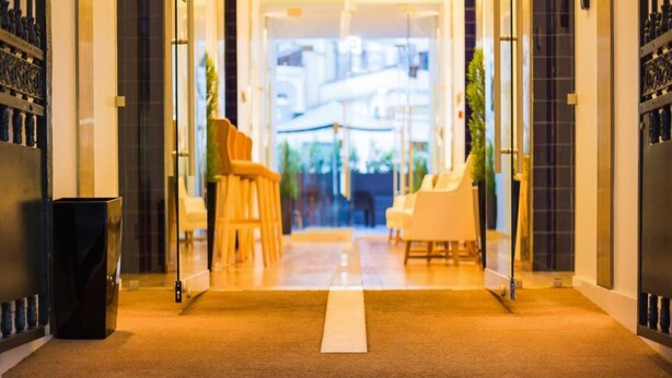 Entrada de un hotel con una alfombra que dice "HOTEL PORTUGAL" y una puerta de vidrio que da a una zona de estar con sillas.