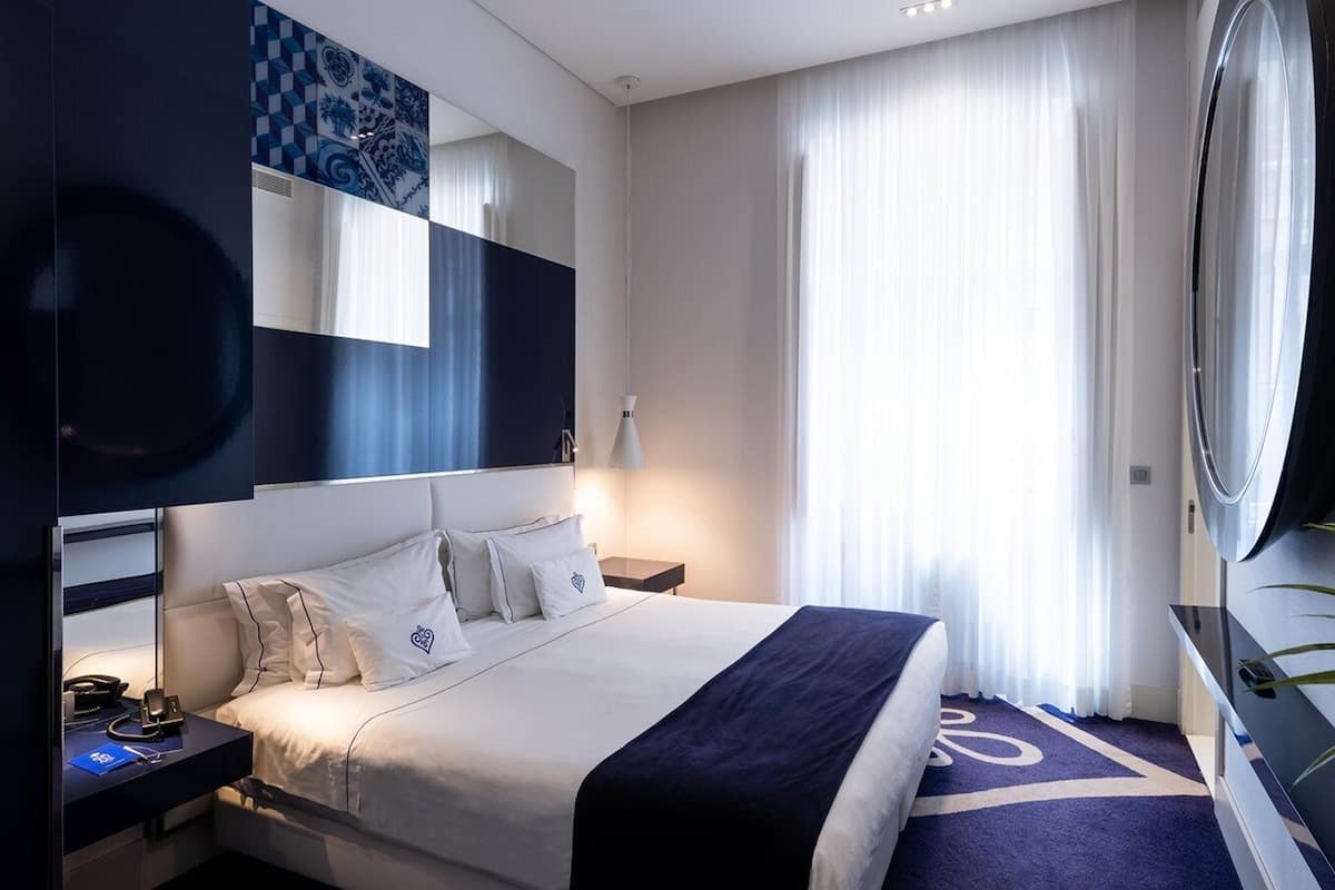 Habitación de hotel moderna con una cama grande, una mesita de noche con teléfono, un espejo y un ventanal con cortinas translúcidas.