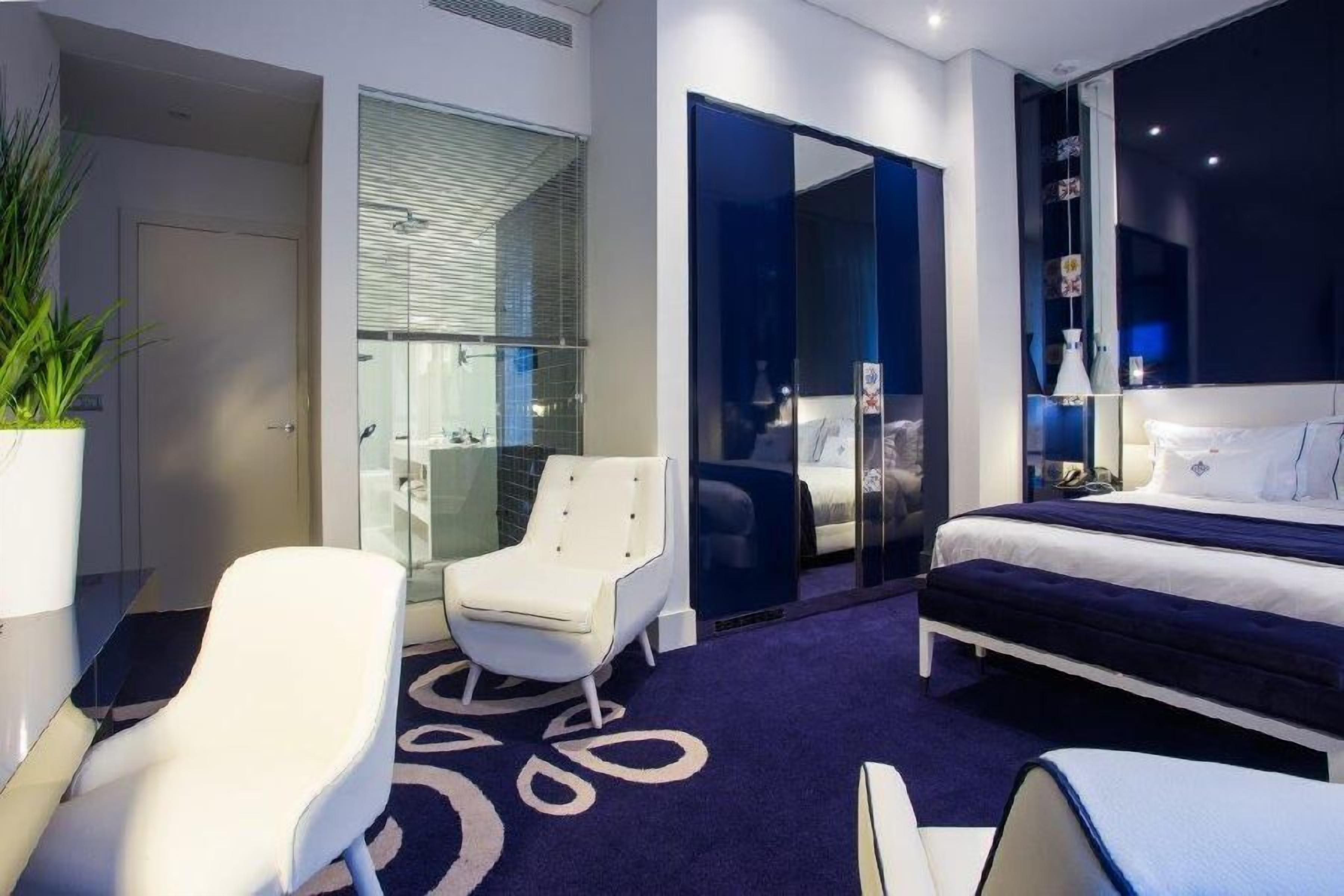 Una habitación de hotel moderna con una cama, un rincón de descanso con una silla, un separador de vidrio y una planta en maceta.