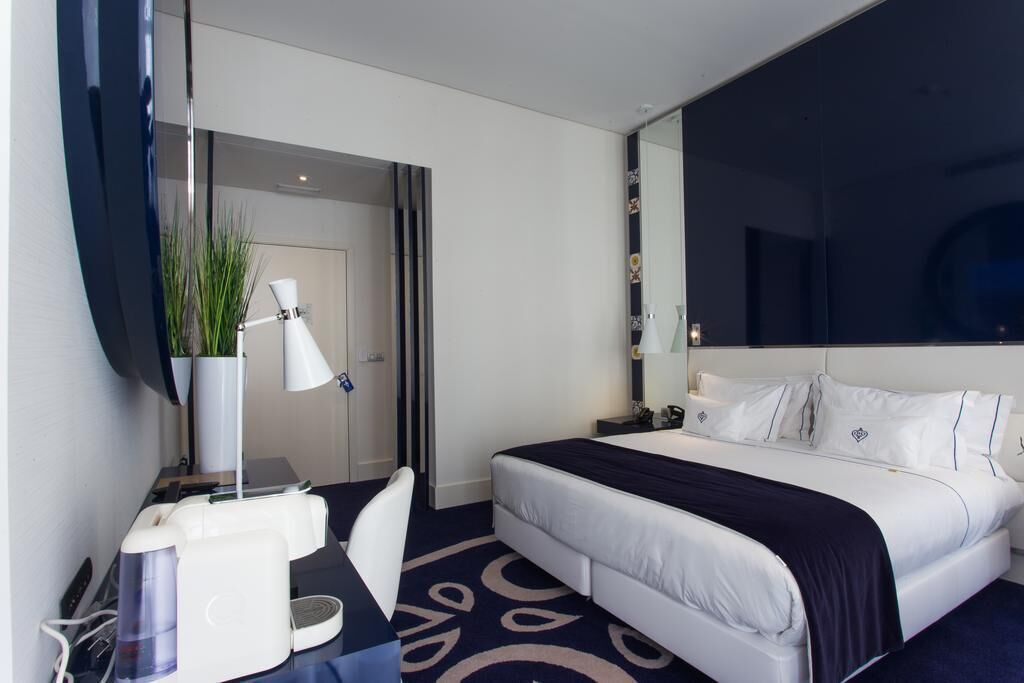 Una habitación de hotel con cama, escritorio con lámpara, silla, espejo grande y planta en maceta.
