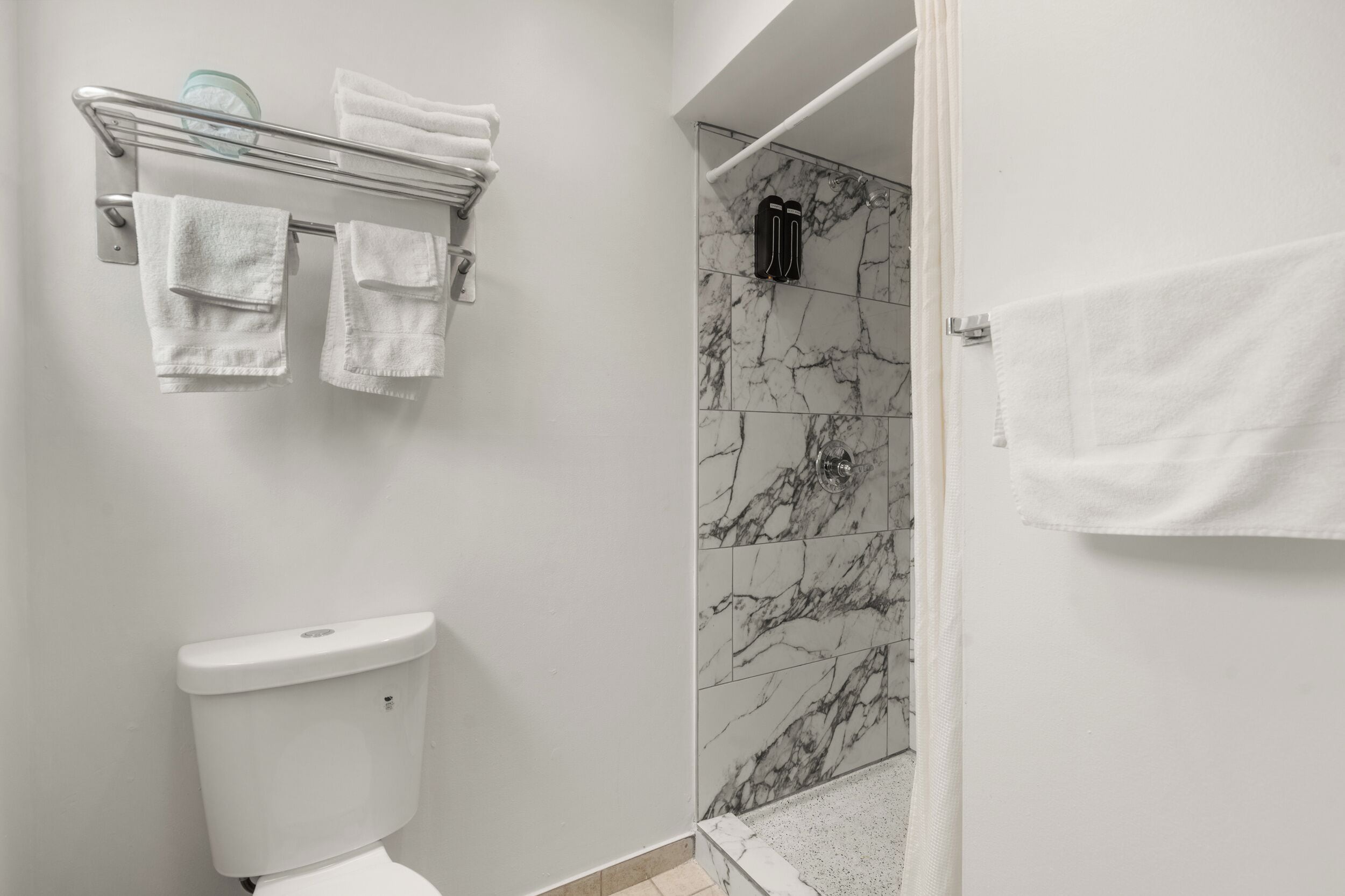 A bathroom with a toilet, a shower with marble tiles, and a towel rack with folded towels.