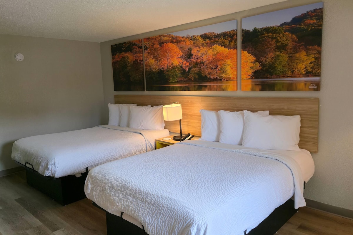 A hotel room with two beds, a nightstand with a lamp, and a large autumn-themed painting on the wall.