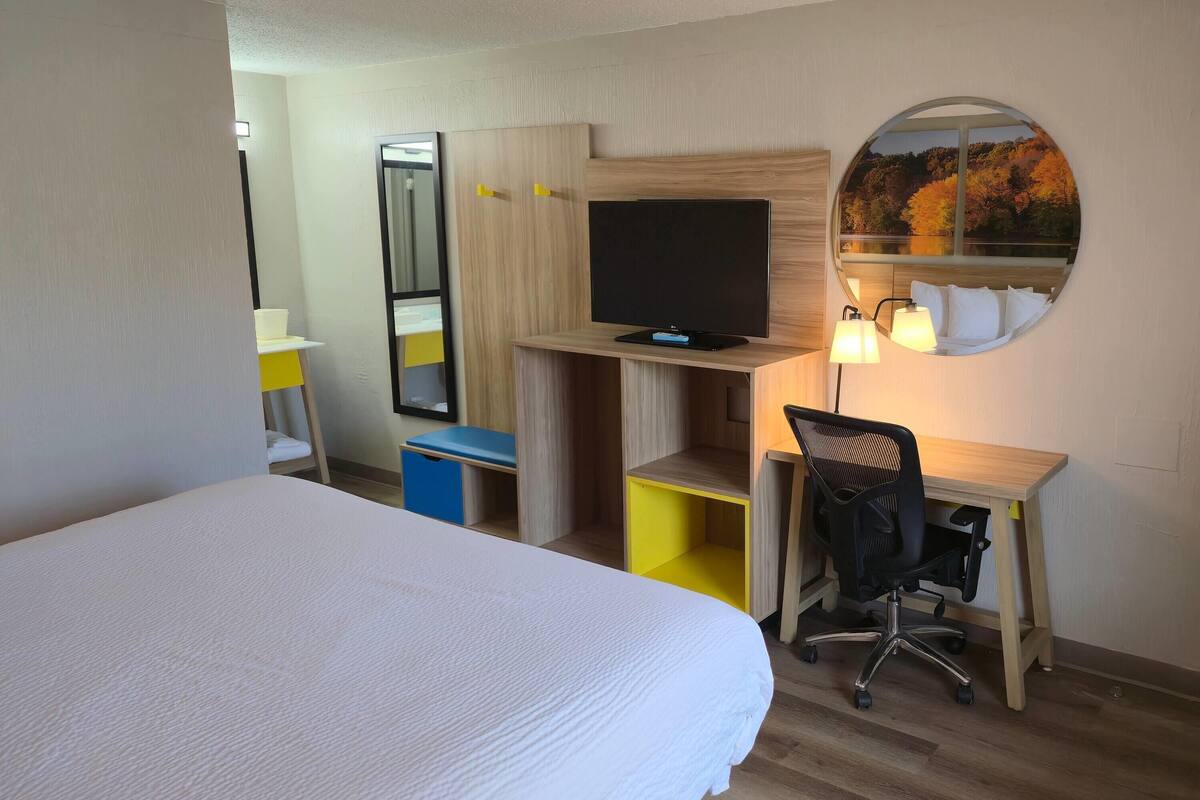A hotel room with a bed, a desk with a computer, a TV, and a mirror with a scenic view.