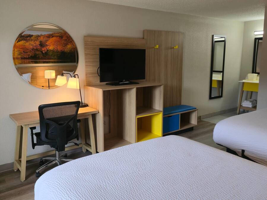 A hotel room with a bed, a desk with a chair, a television, a mirror, and a storage unit.