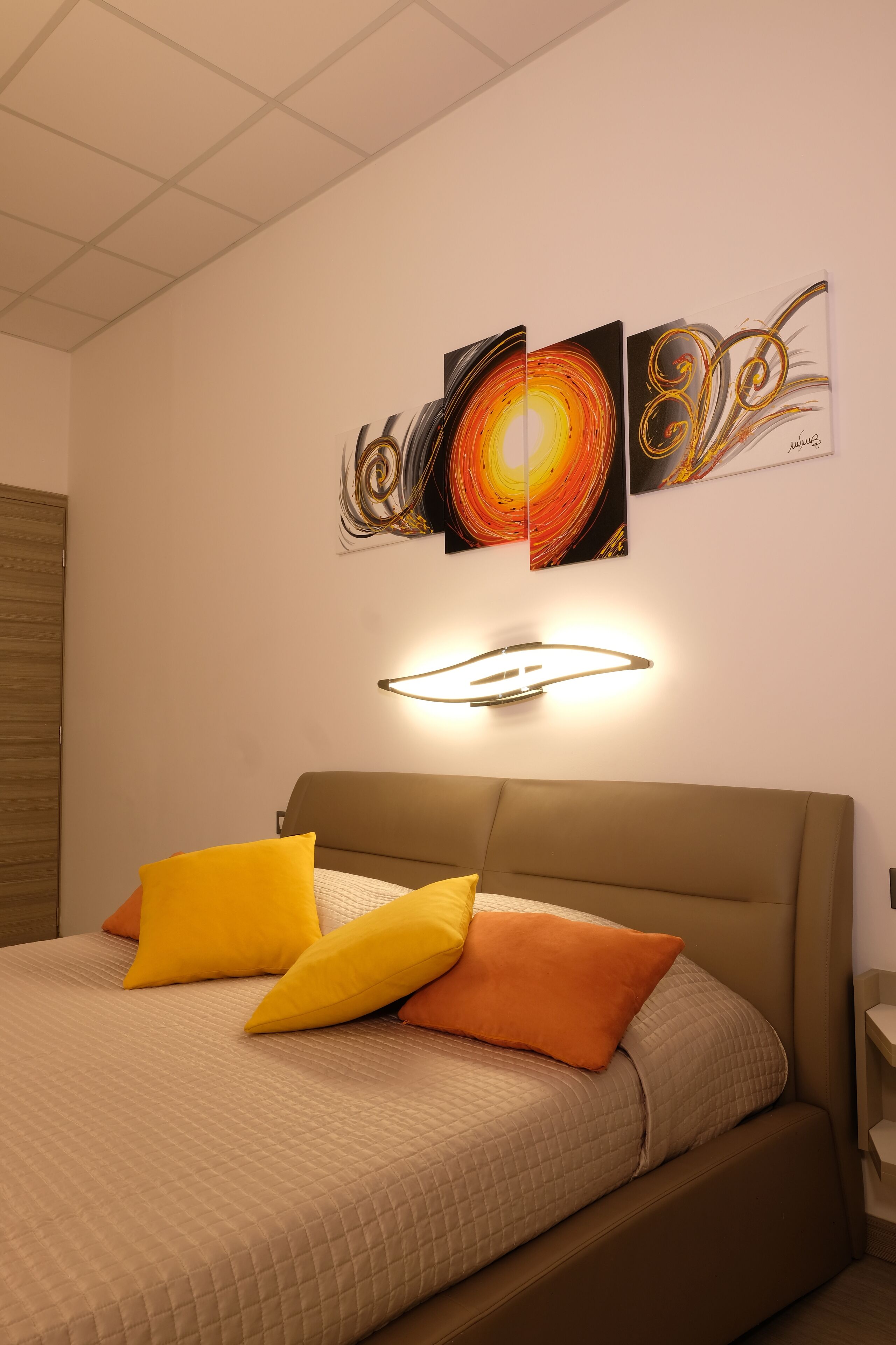 A bedroom with a bed, two pillows, a wall-mounted light, and three pieces of wall art.
