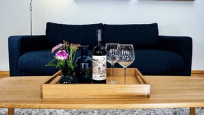 Une table basse en bois, avec une bouteille de vin, deux verres à vin et un vase de fleurs, devant un canapé bleu foncé.