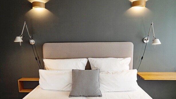 Un lit avec une tête de lit grise, des oreillers blancs et gris, et deux lampes fixées au mur de chaque côté.