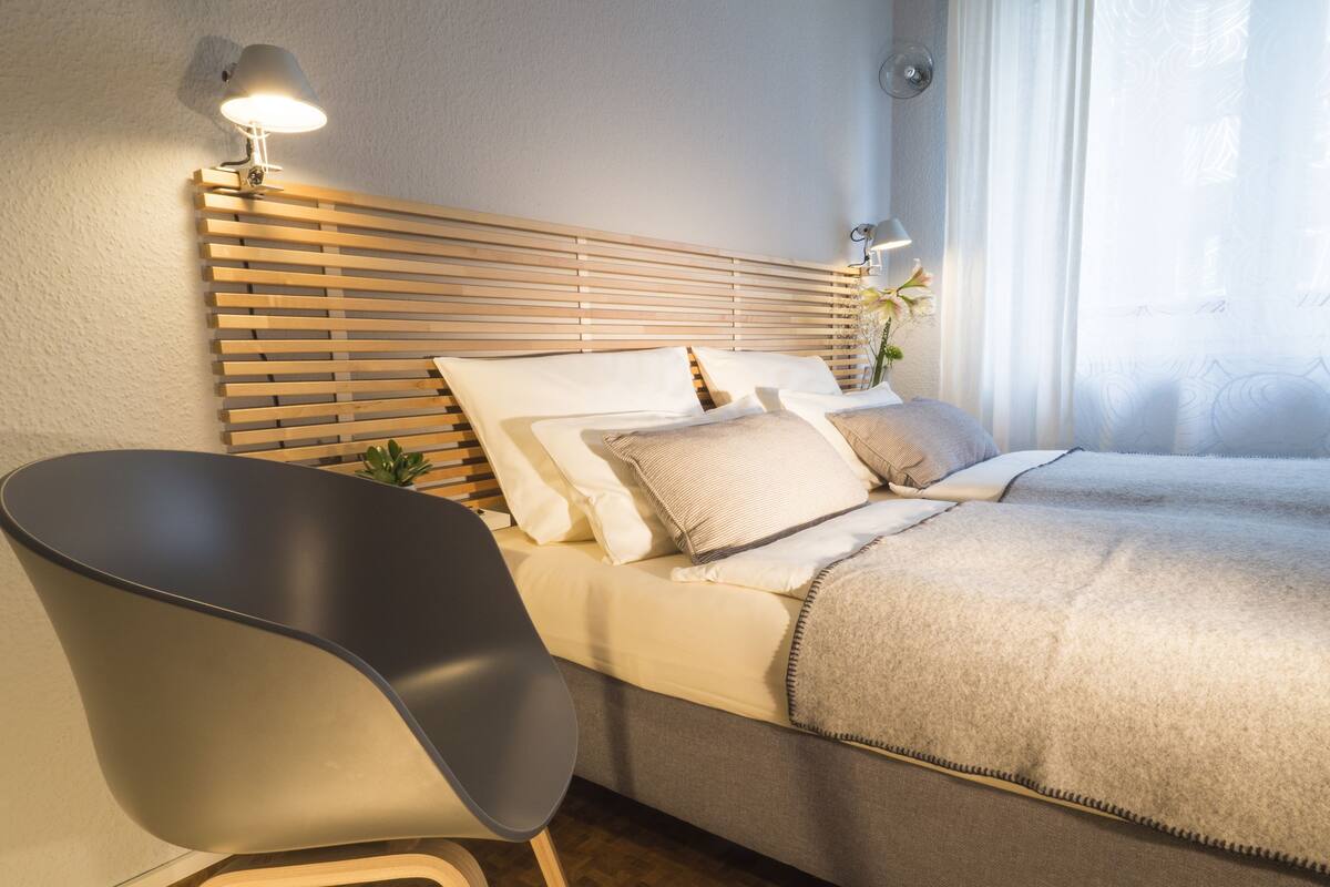 A modern bedroom with a wooden slat headboard, a bed with multiple pillows, a grey chair, and a bedside table with a lamp.